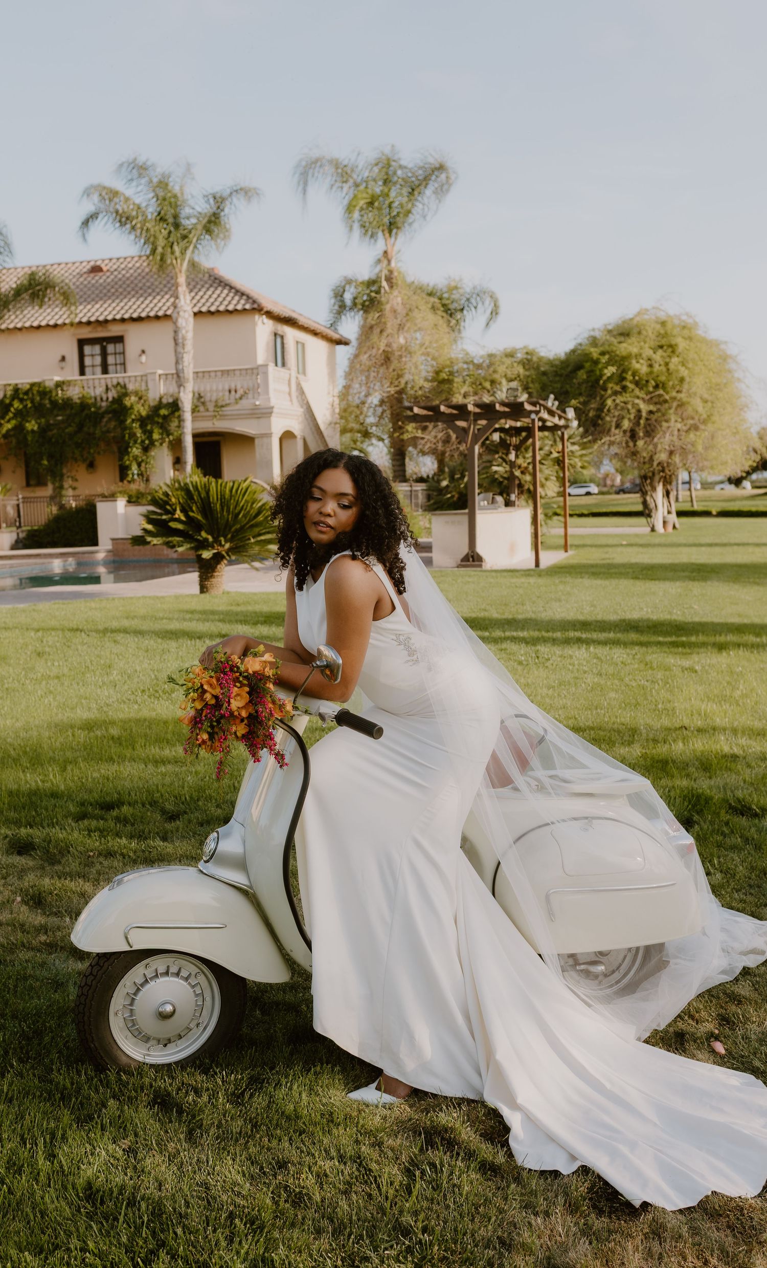 Bride in white gown with veil poses on a white vintage Vespa scooter holding a vibrant bouquet at sunset.