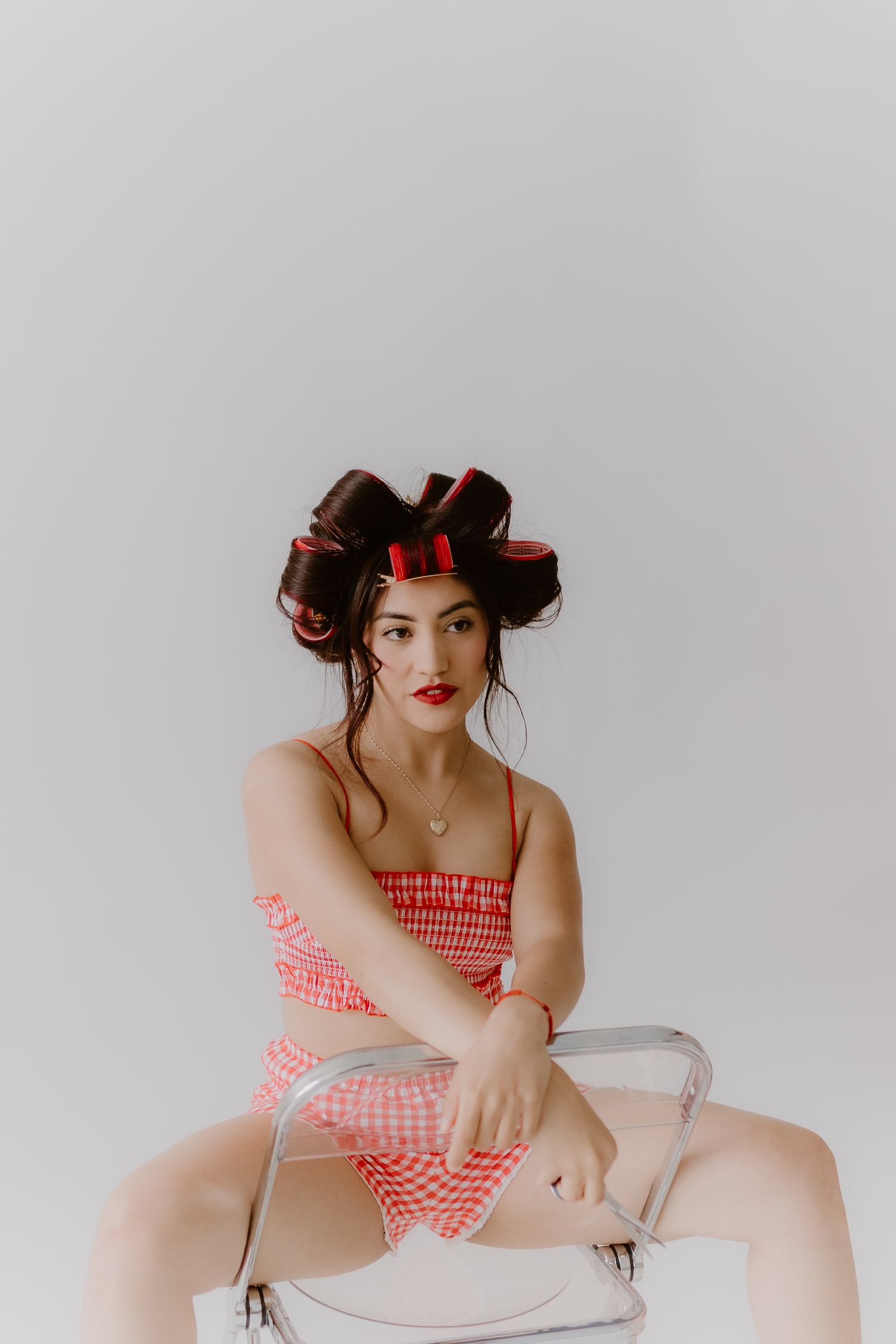 Young woman in red gingham dress with hair rollers sits on clear chair against white background.