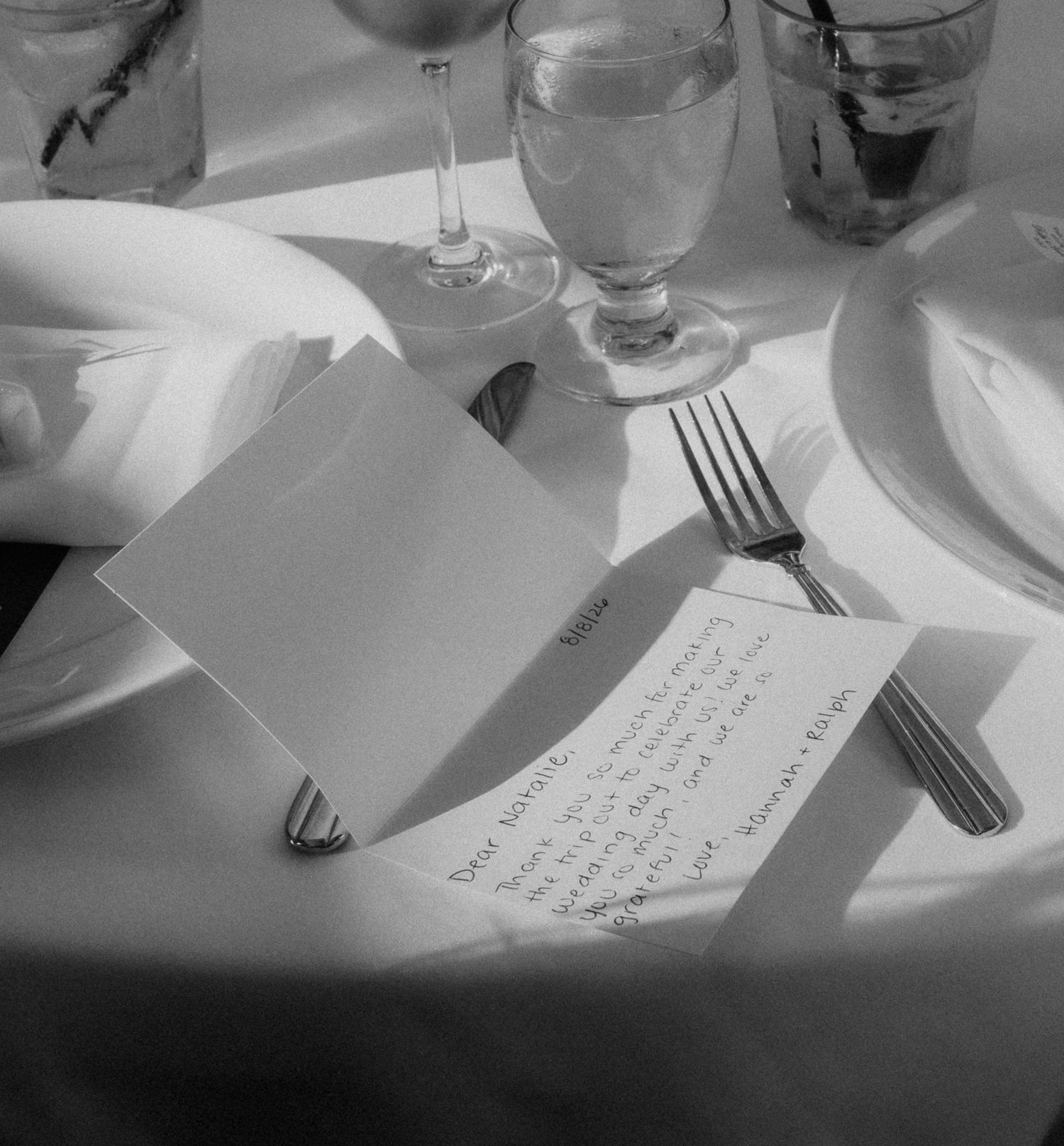 Black and white close-up of handwritten wedding place card beside fork and glassware on reception table.