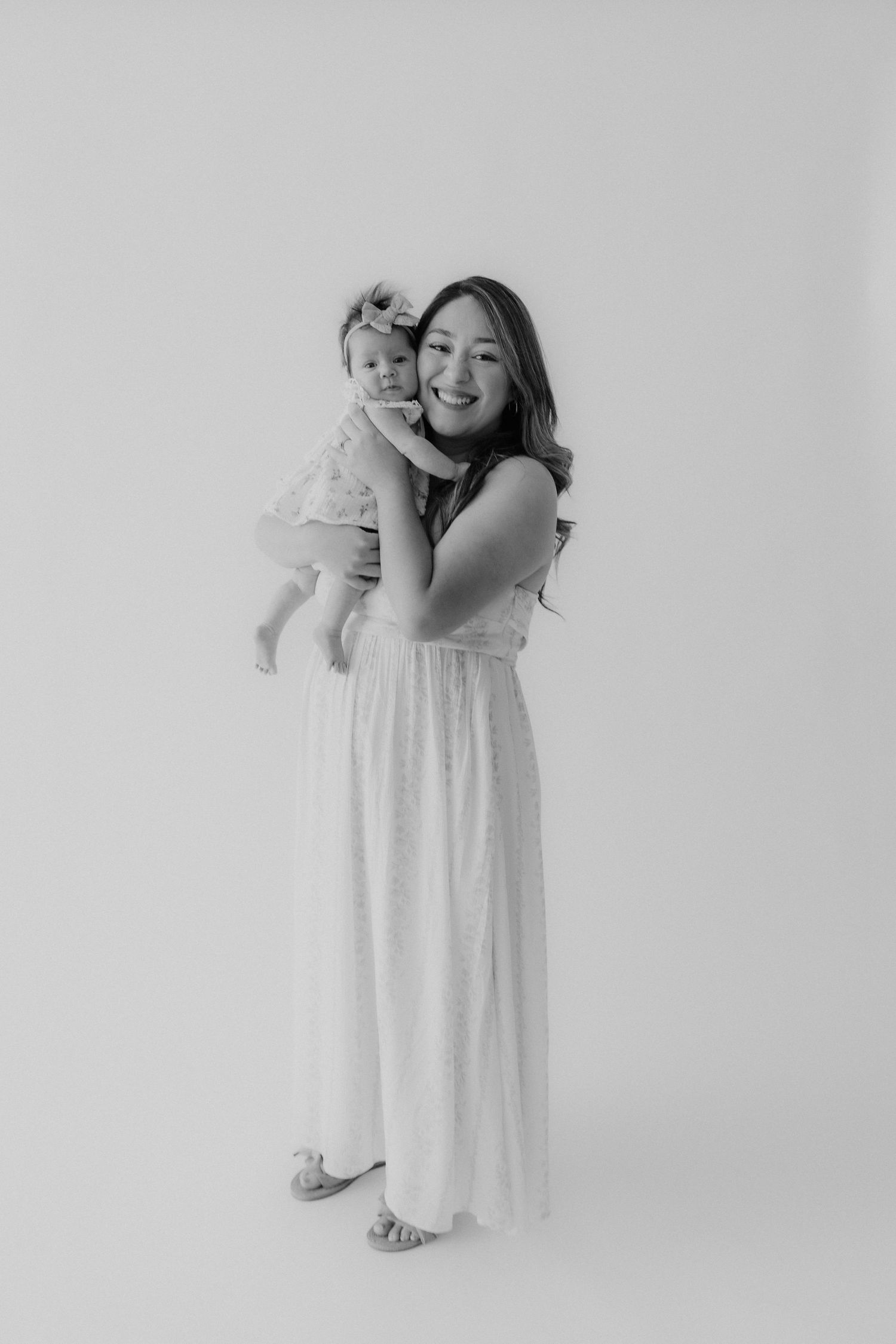 Full-length black and white photo of a young woman in a white dress holding a smiling baby girl with a headband.