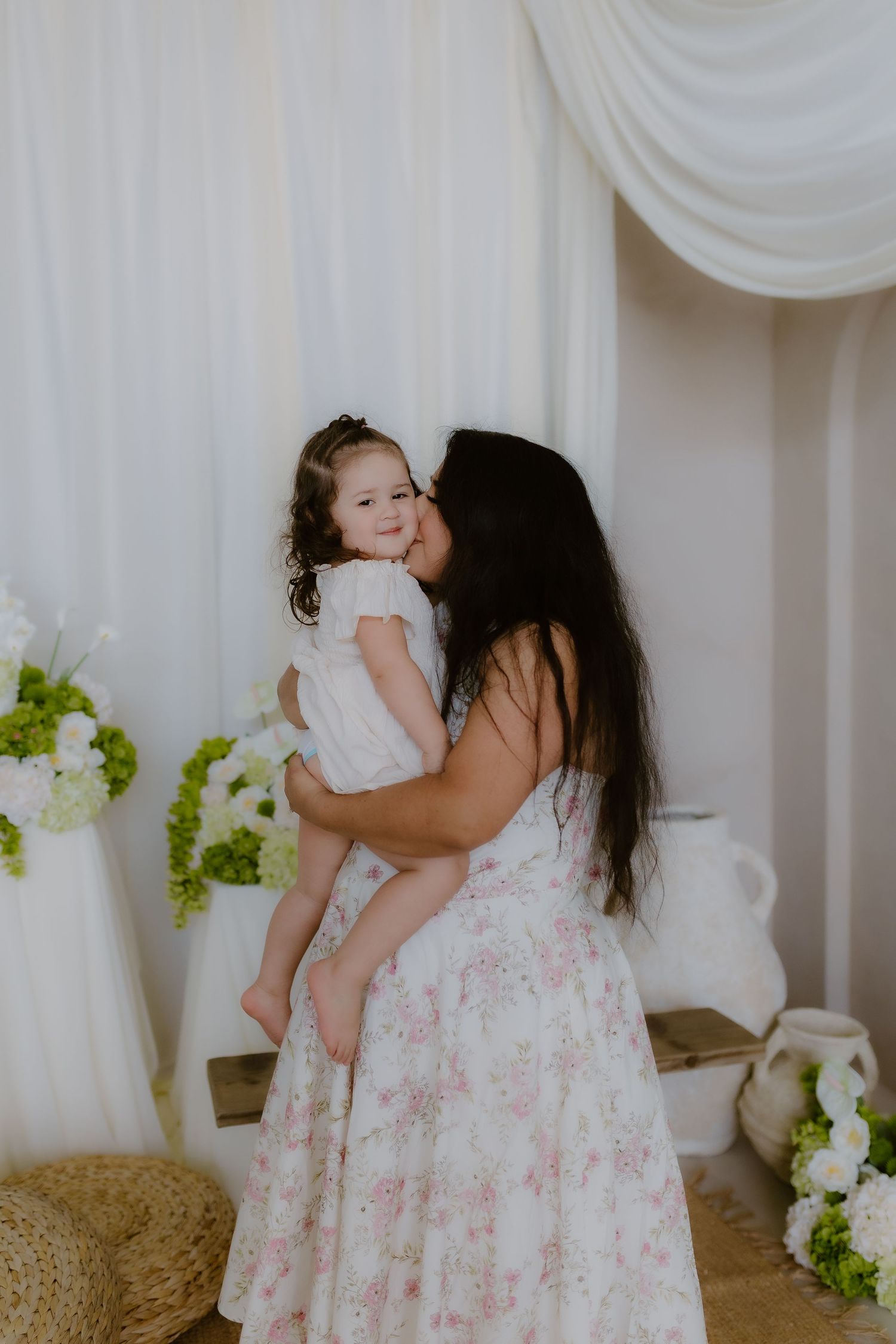 Mother in floral dress kissing toddler daughter in white dress in elegant white studio setting.
