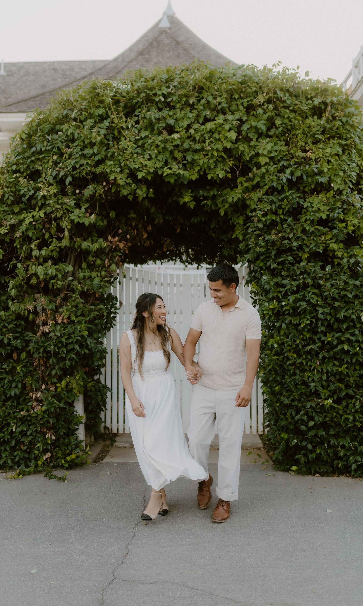 Couple in white outfits holding hands and walking through ivy-covered garden gate.