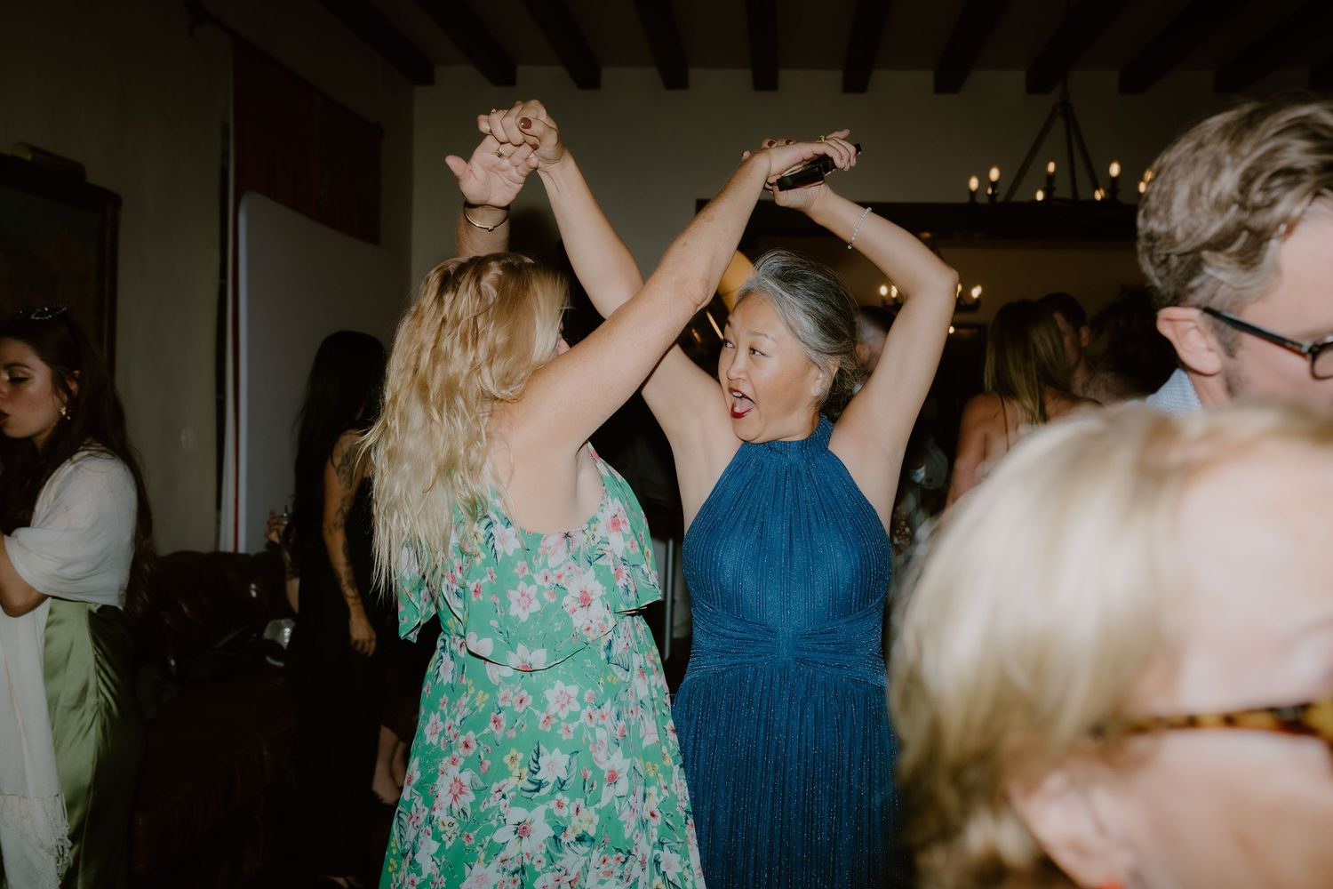 Wedding guests in floral and teal dresses dancing together at indoor reception celebration.