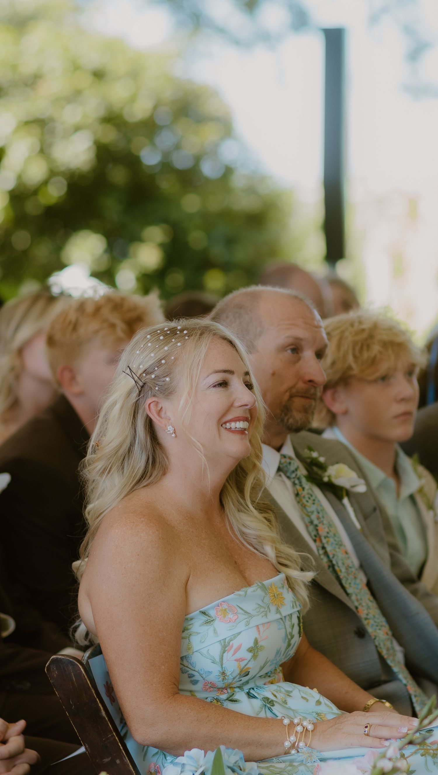 Smiling wedding guest in a floral dress enjoys an outdoor ceremony seated among other guests.