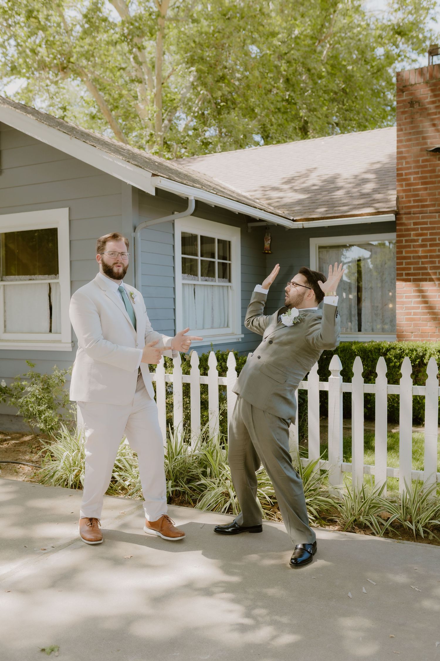 Two groomsmen in suits celebrating with playful high-five gesture outside blue house.