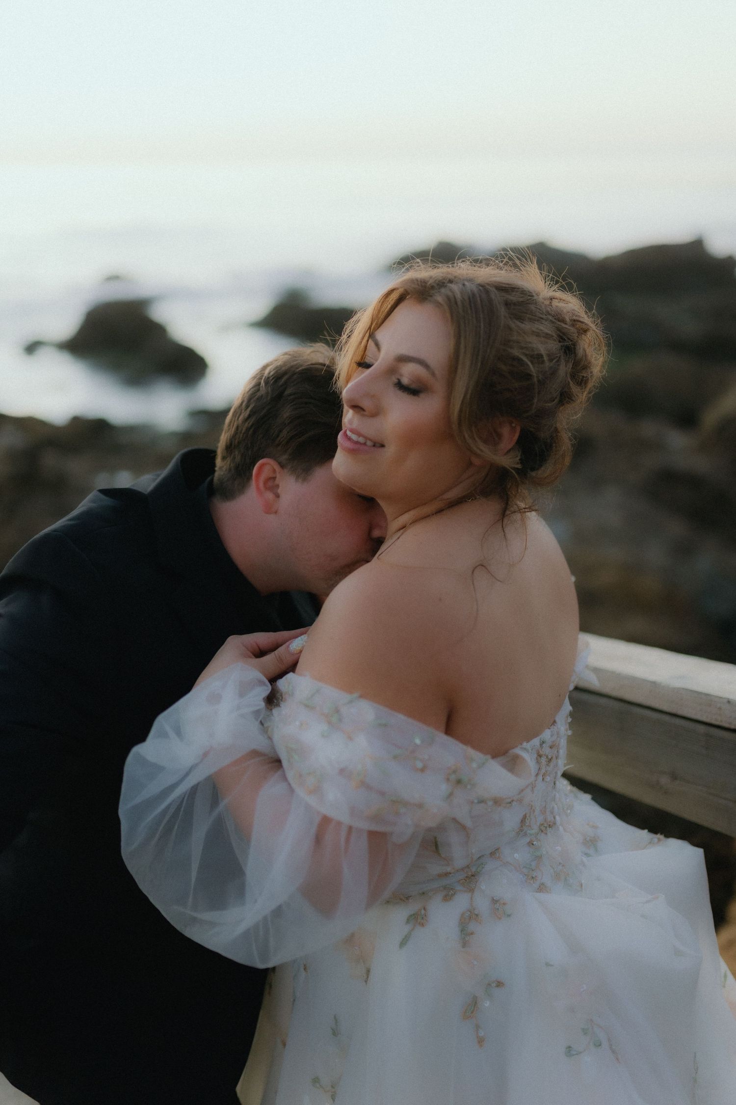 Groom kissing bride on shoulder at rocky ocean overlook at sunset.
