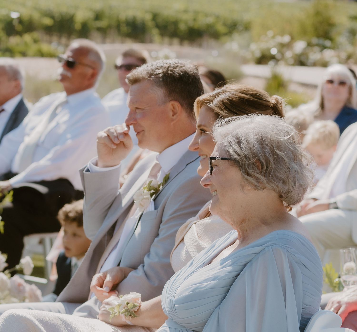 Wedding guests smiling and laughing during an outdoor ceremony at a vineyard on a sunny day.