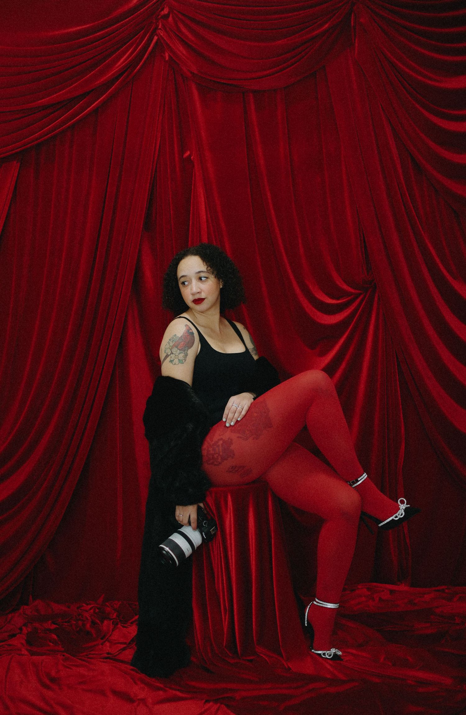 Female photographer in black outfit and red tights posing with camera against red velvet curtains.