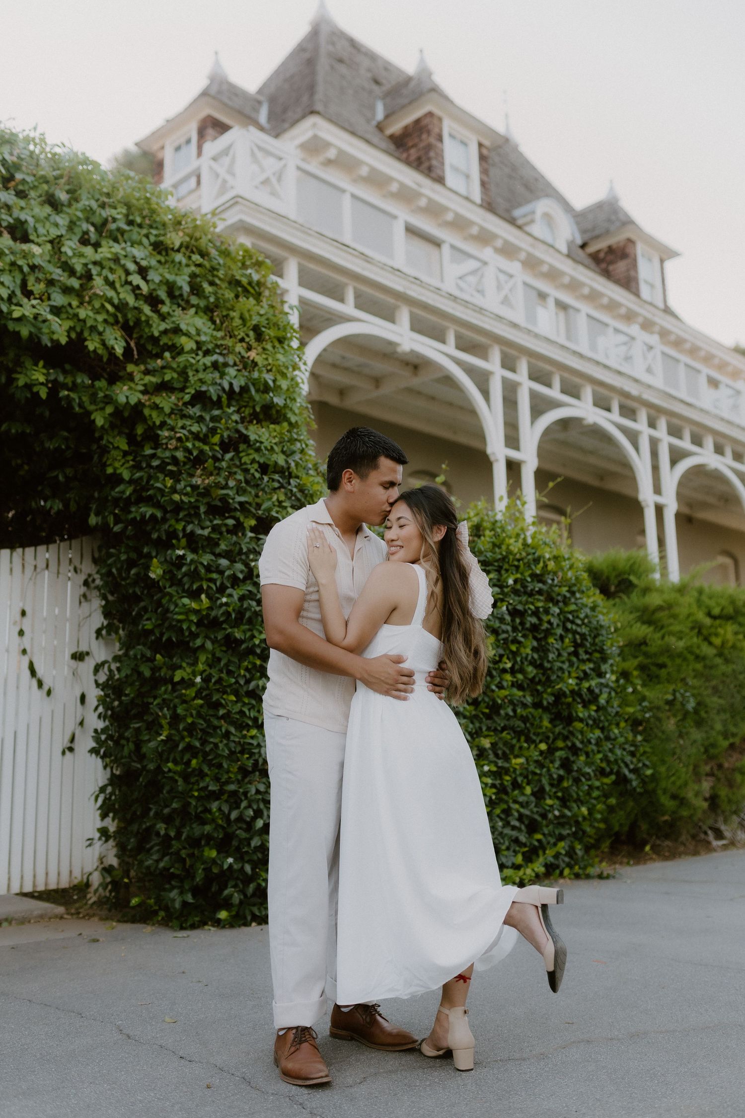 Couple in white outfits embracing in front of a charming ivy-covered Victorian mansion at sunset.