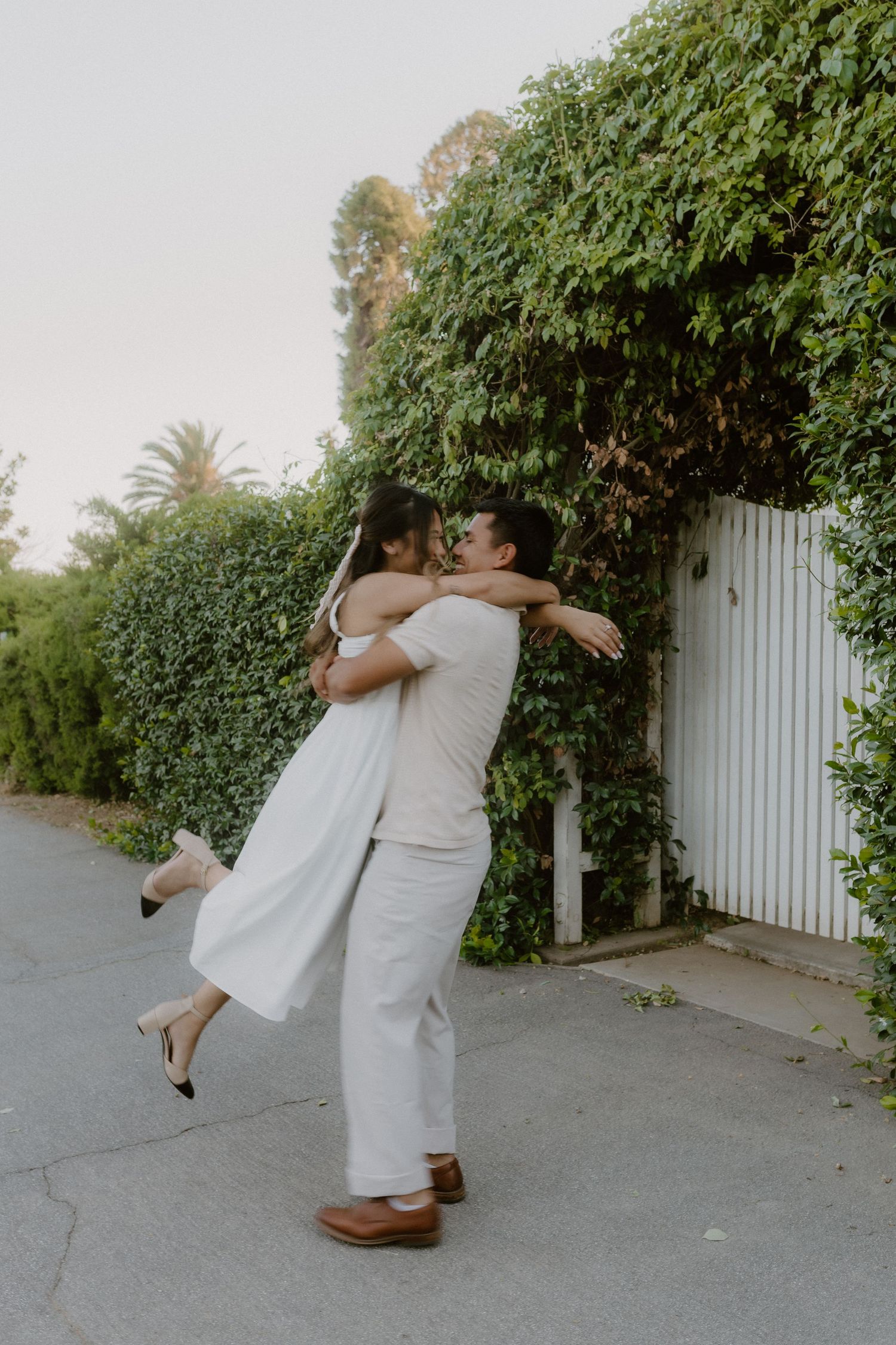 Man lifting woman in white dress while embracing near an ivy-covered white gate.