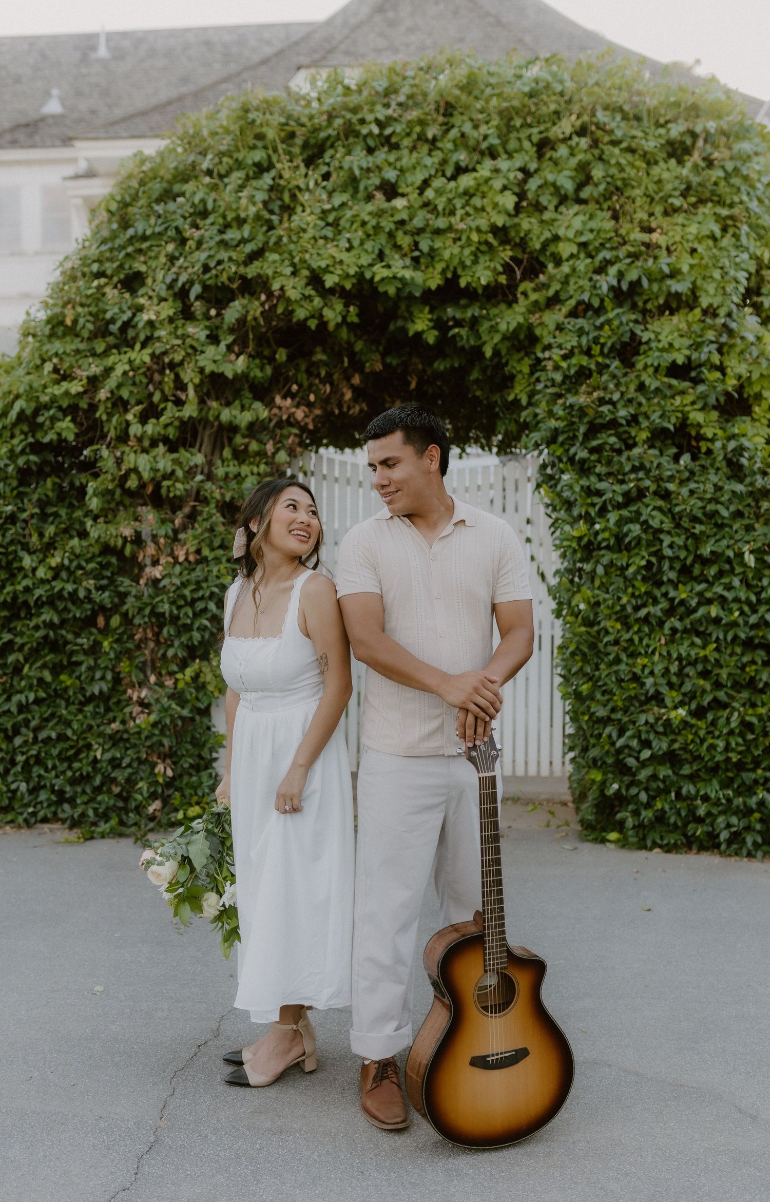 Couple in white outfits standing together near ivy-covered arch with acoustic guitar outdoors.