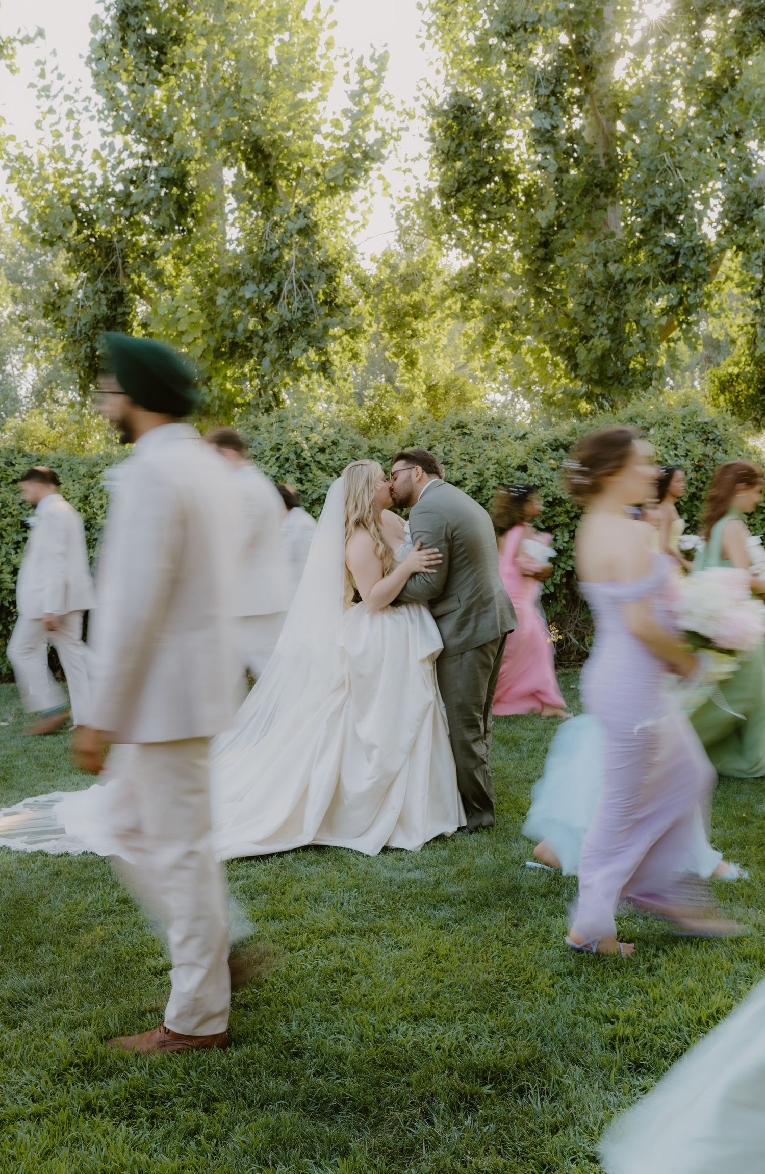 Bride and groom sharing intimate kiss surrounded by moving wedding guests in garden setting.