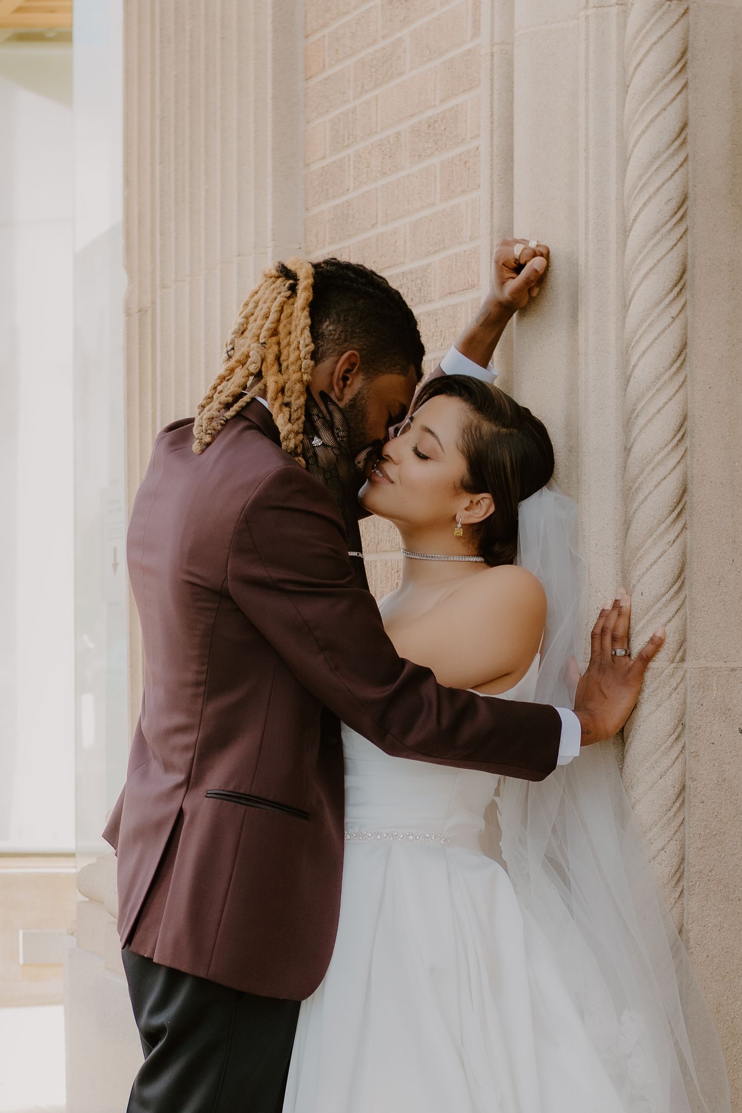 Groom in burgundy suit with dreadlocks kissing bride in white gown against ornate stone column at wedding venue.
