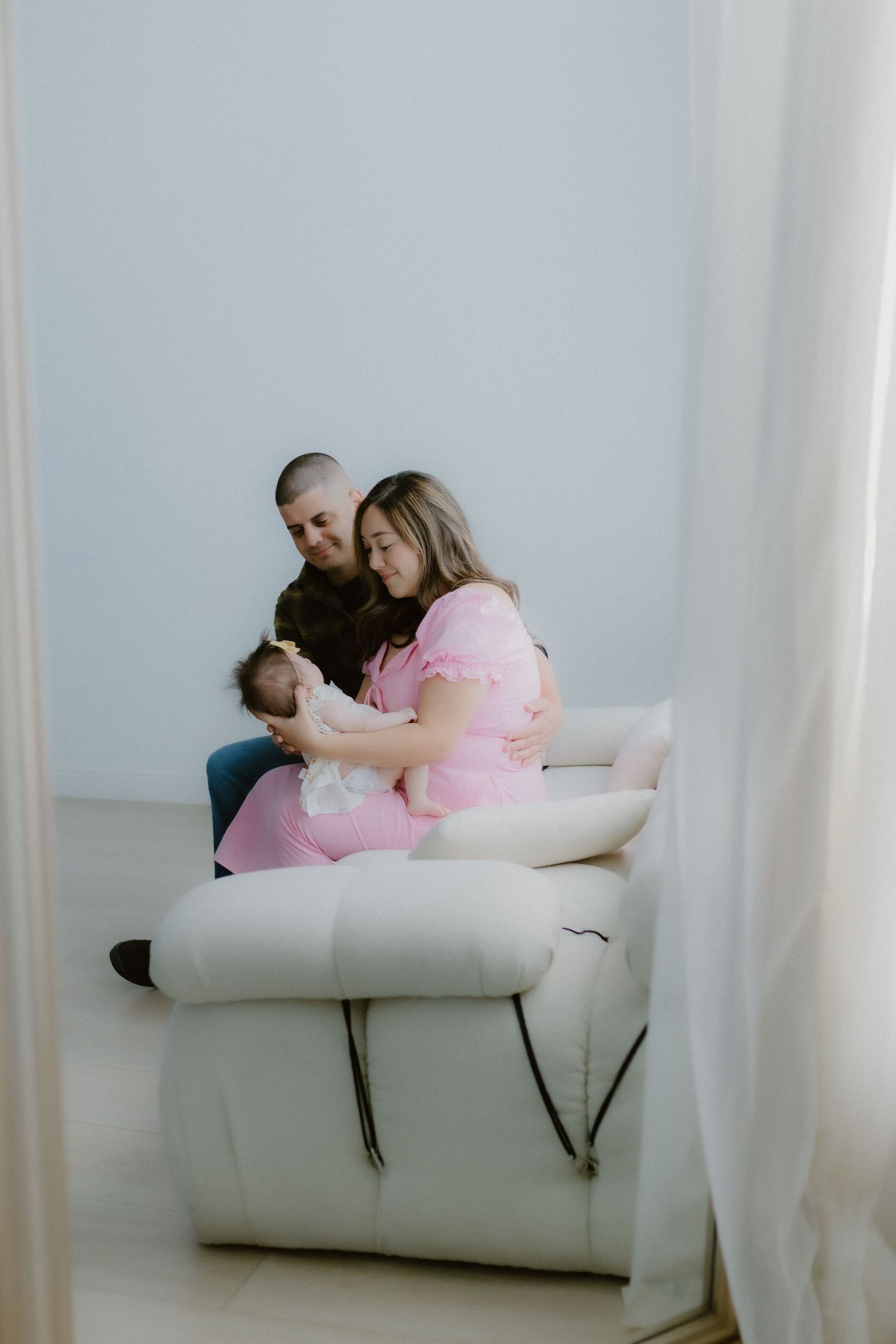 Young couple in pink and plaid sitting on white leather sofa cradling their newborn baby.