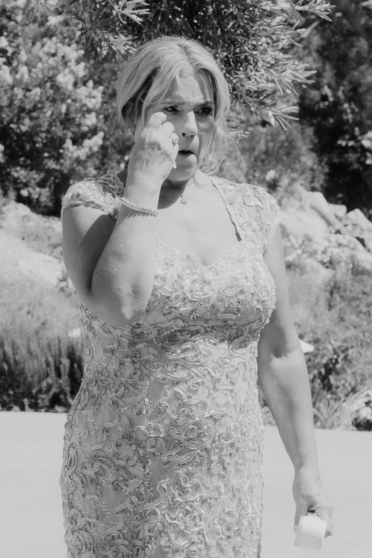 Emotional woman in lace dress wiping tears at an outdoor wedding ceremony in black and white.