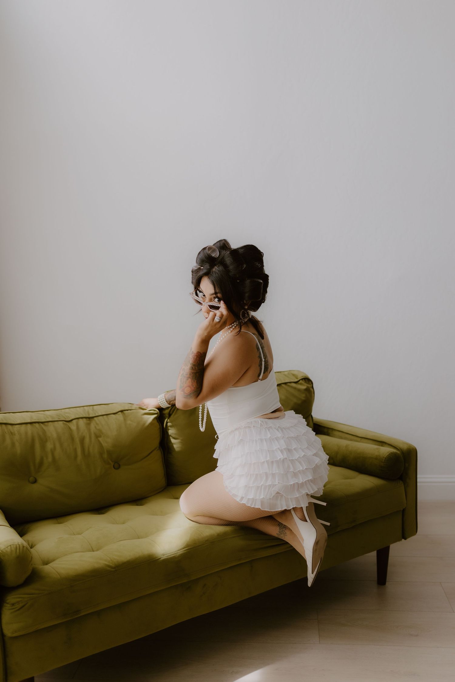 Woman in white ruffled mini dress kneeling on green velvet sofa in bright minimalist room. Tulare California, birthday