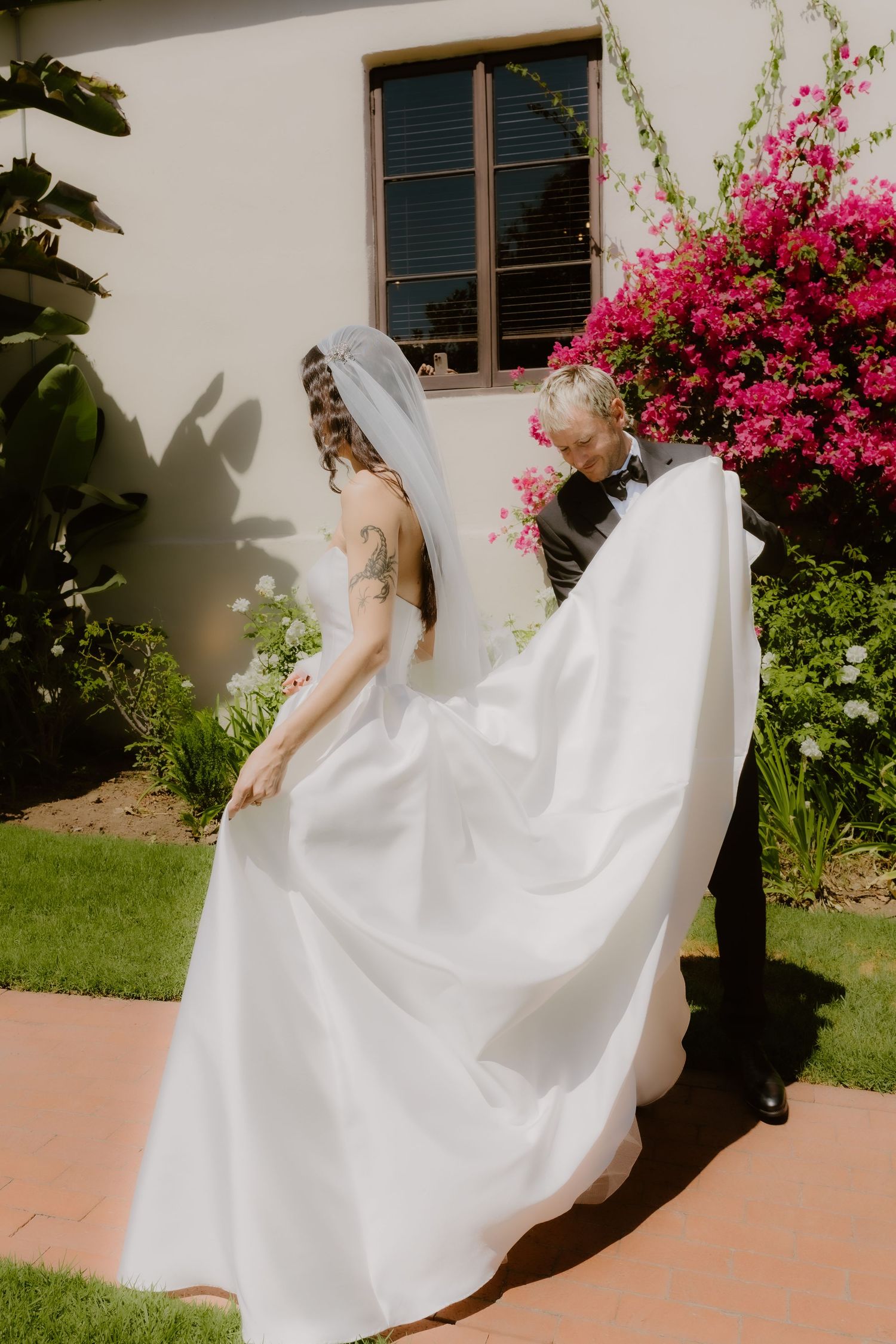 Groom lifting bride's white wedding dress train near blooming pink bougainvillea against white stucco wall.