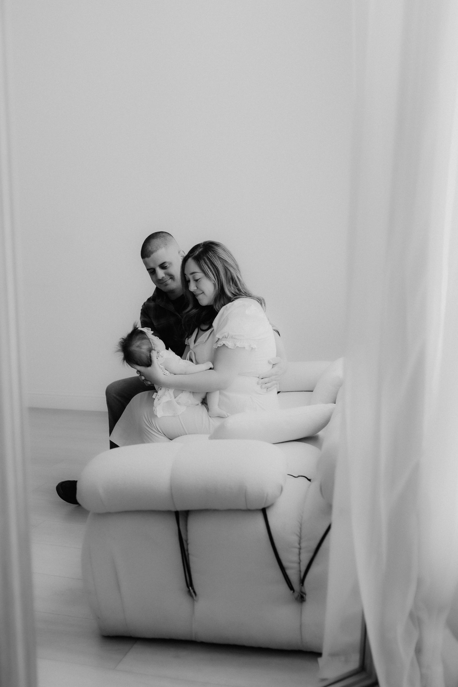 Black and white photo of couple sitting on white sofa lovingly holding their newborn baby.