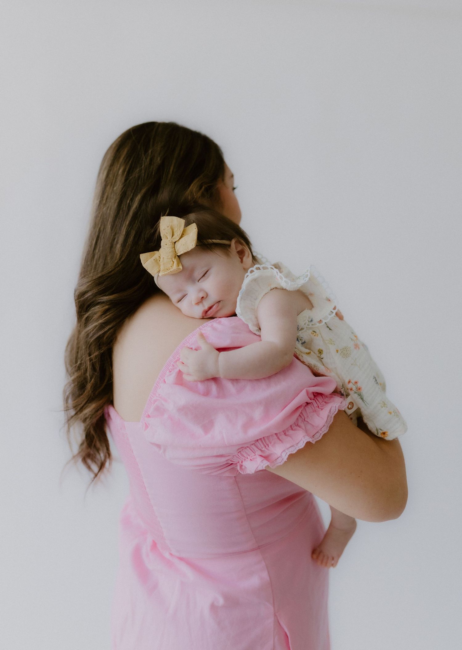 Mother in pink dress holding sleeping baby girl with yellow bow on her shoulder.