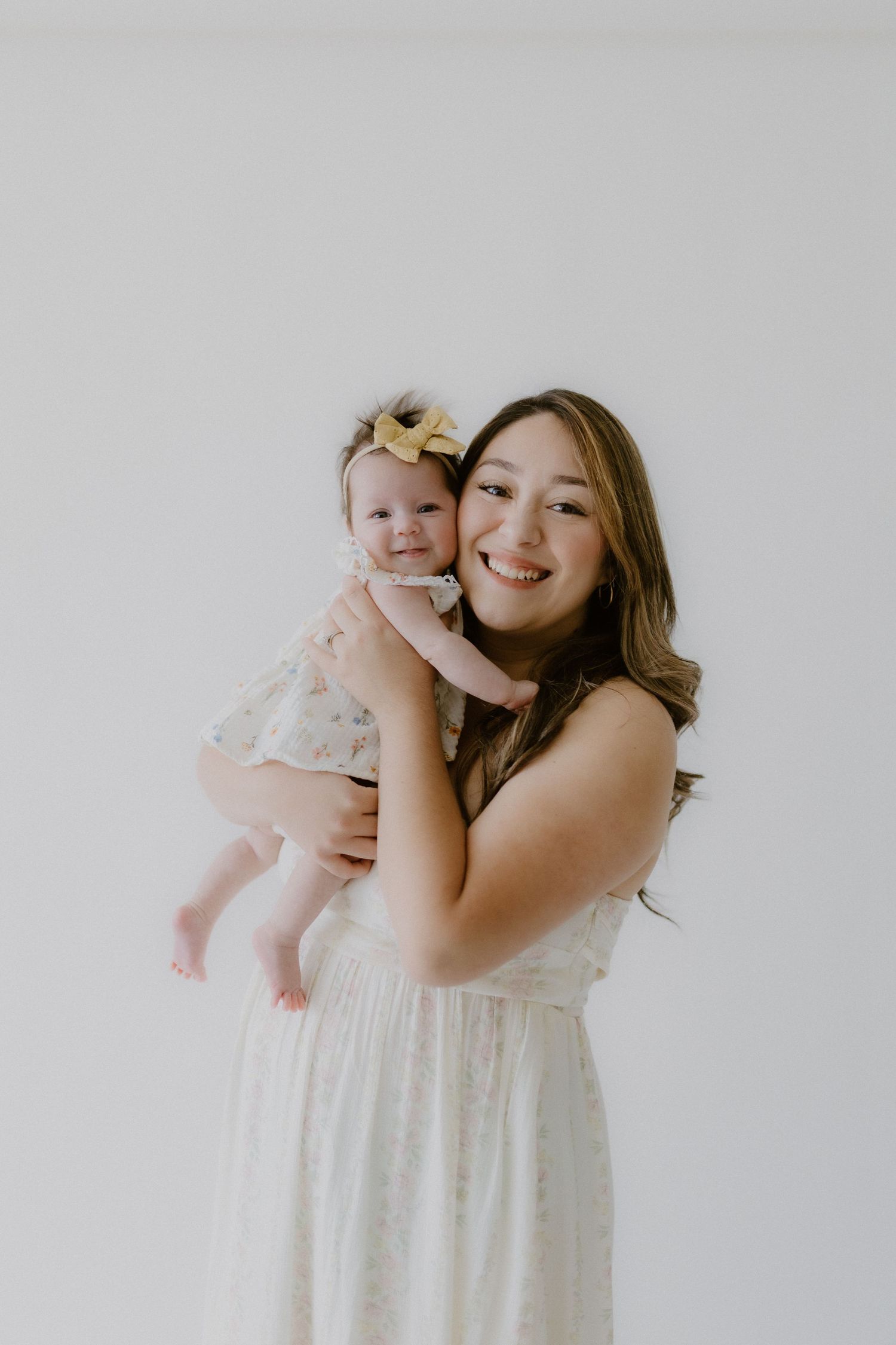 A young woman in a white floral dress smiles while holding a baby girl with a yellow bow in a bright studio.