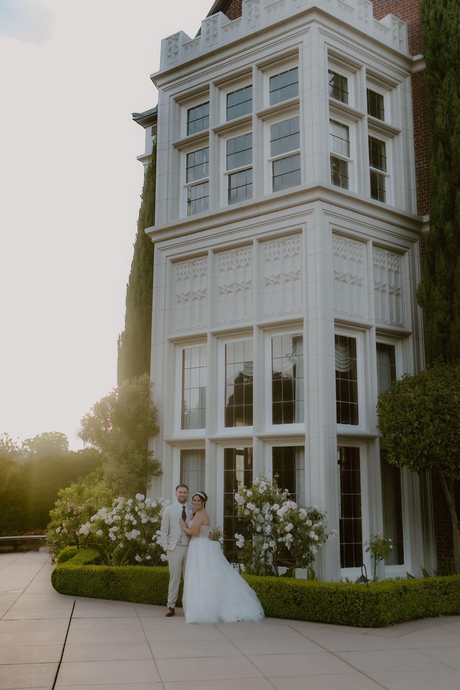 Bride and groom pose at golden hour beside tall ornate white bay window of elegant venue.