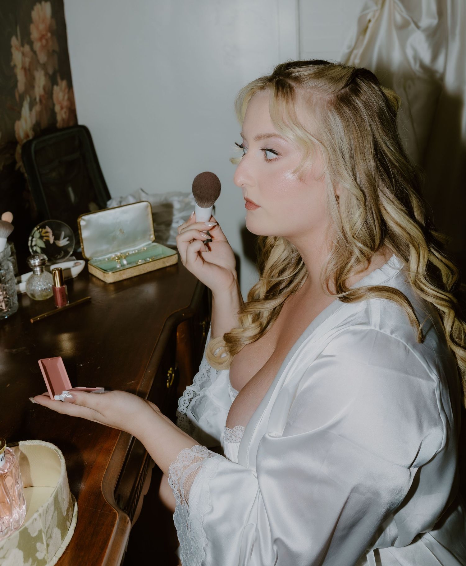 Blonde bride in white robe applying makeup with powder brush at vintage vanity table.