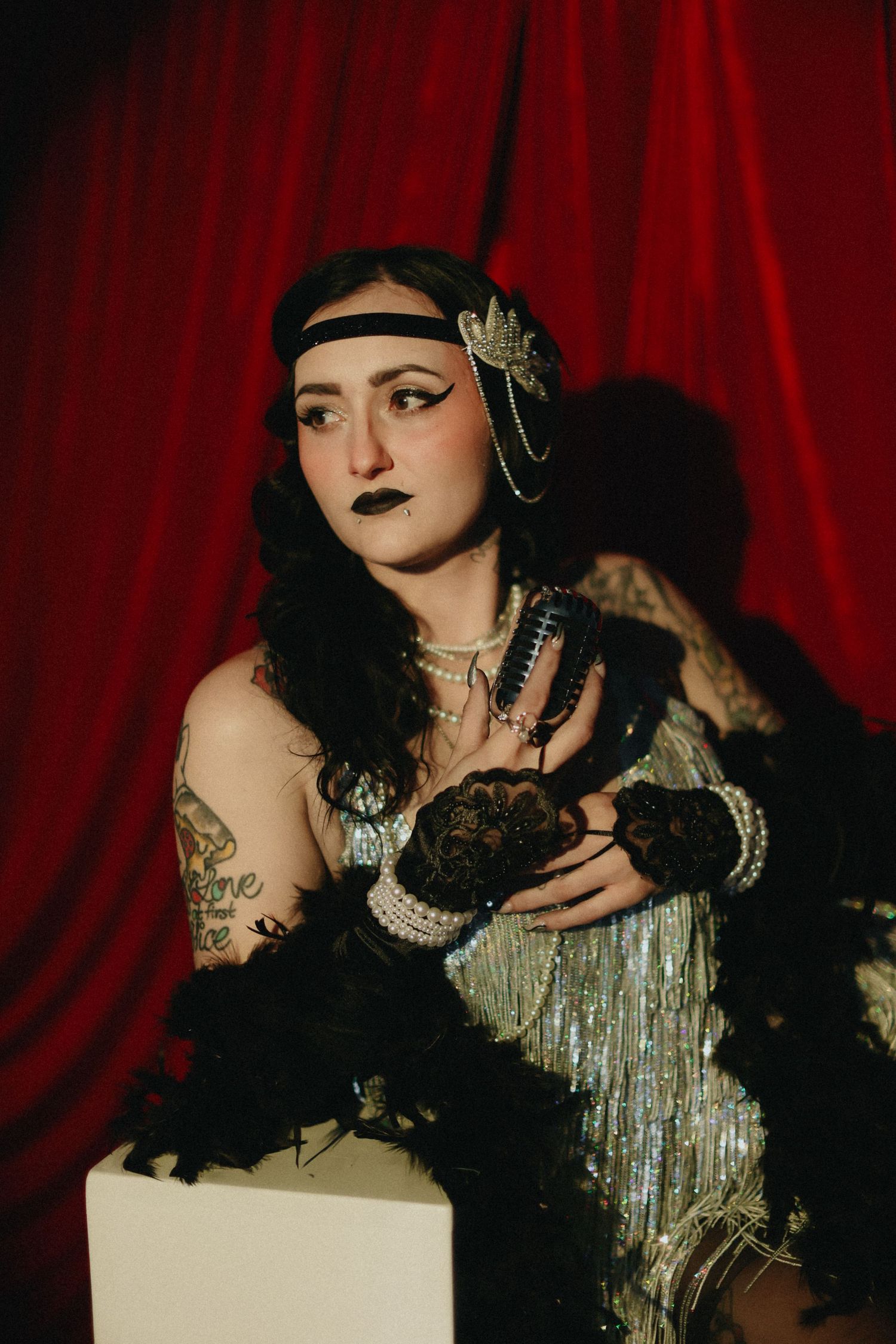 Woman in 1920s flapper costume with black headband, lace gloves, and pearl necklace poses before red curtain.