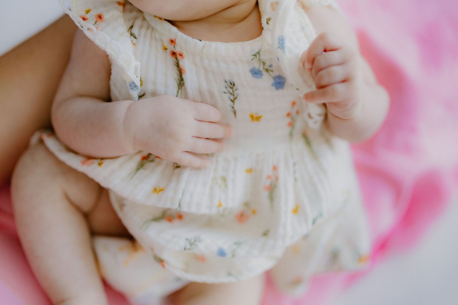 Close-up of a newborn baby in a white floral romper with tiny hands resting on tummy.