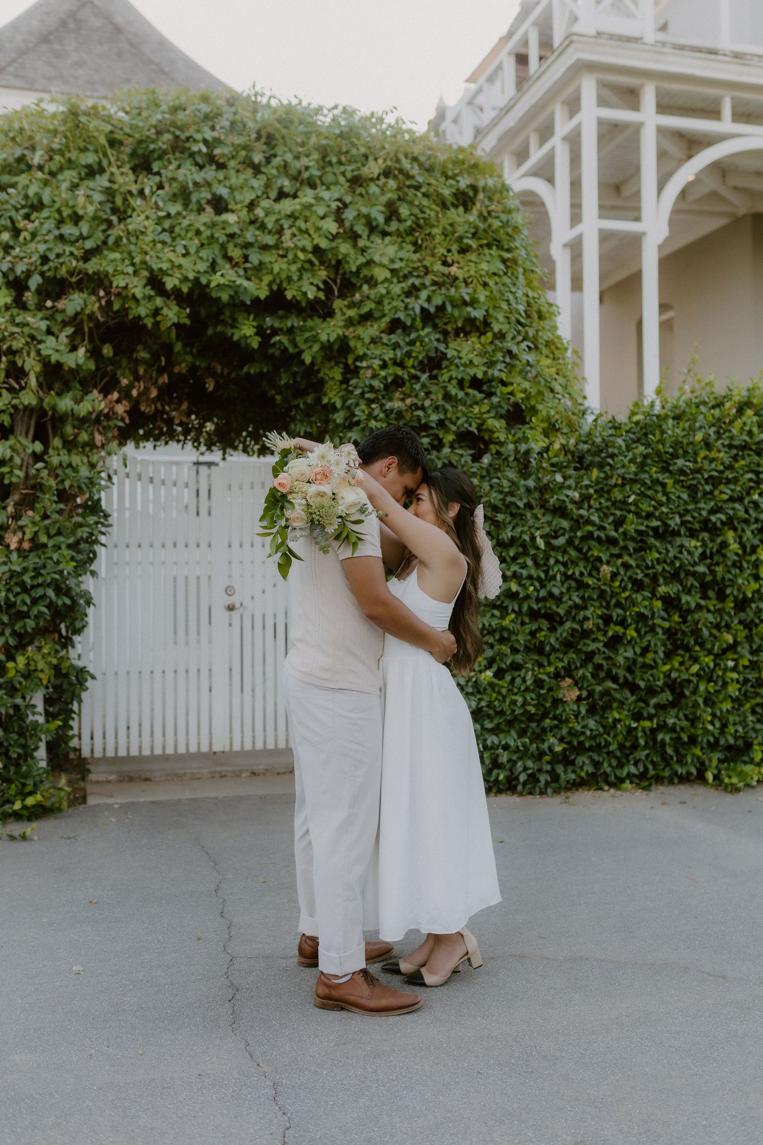 Couple sharing a kiss hidden behind a lush bridal bouquet in front of a white garden gate.