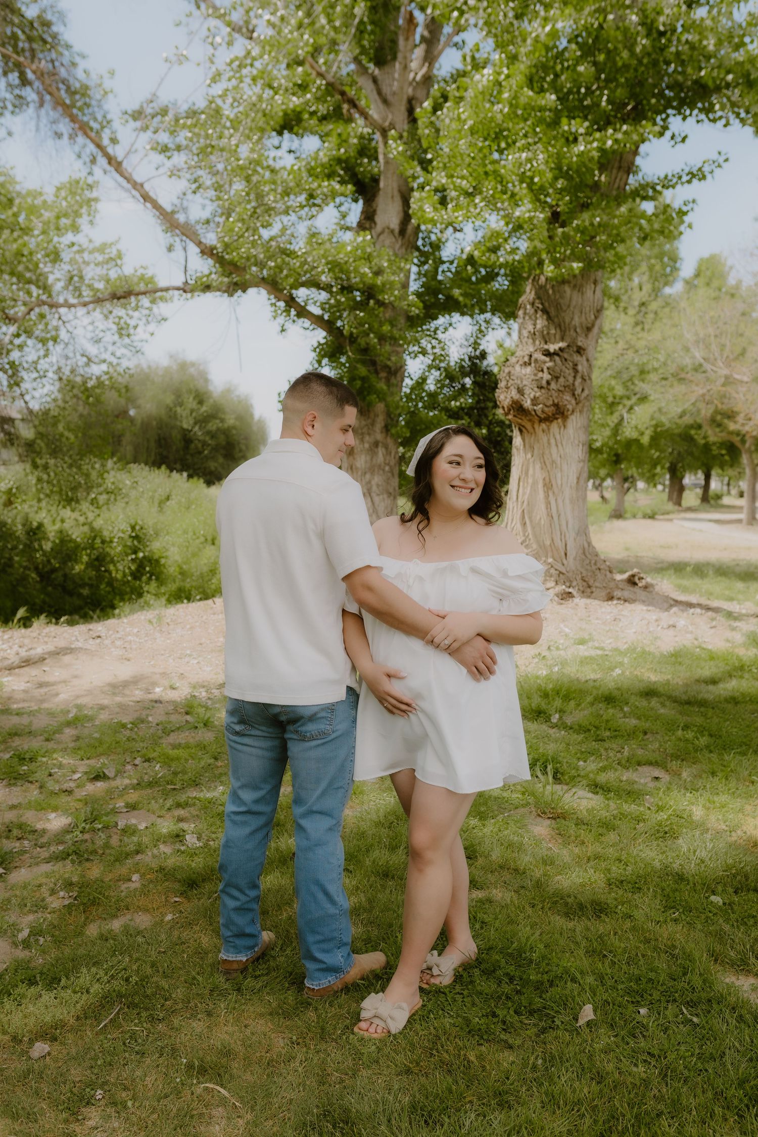Pregnant couple sharing a tender moment outdoors under a large tree, woman in white off-shoulder dress.