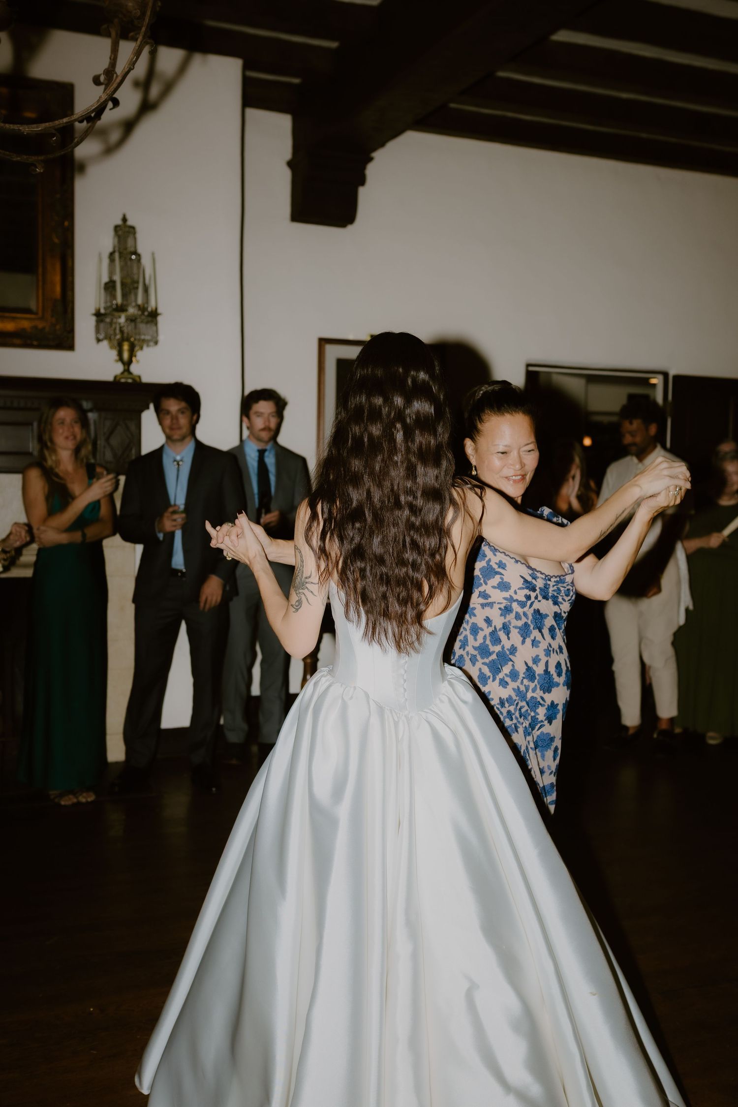 Bride dances joyfully with a female guest in a blue floral dress on the dance floor at the wedding reception.