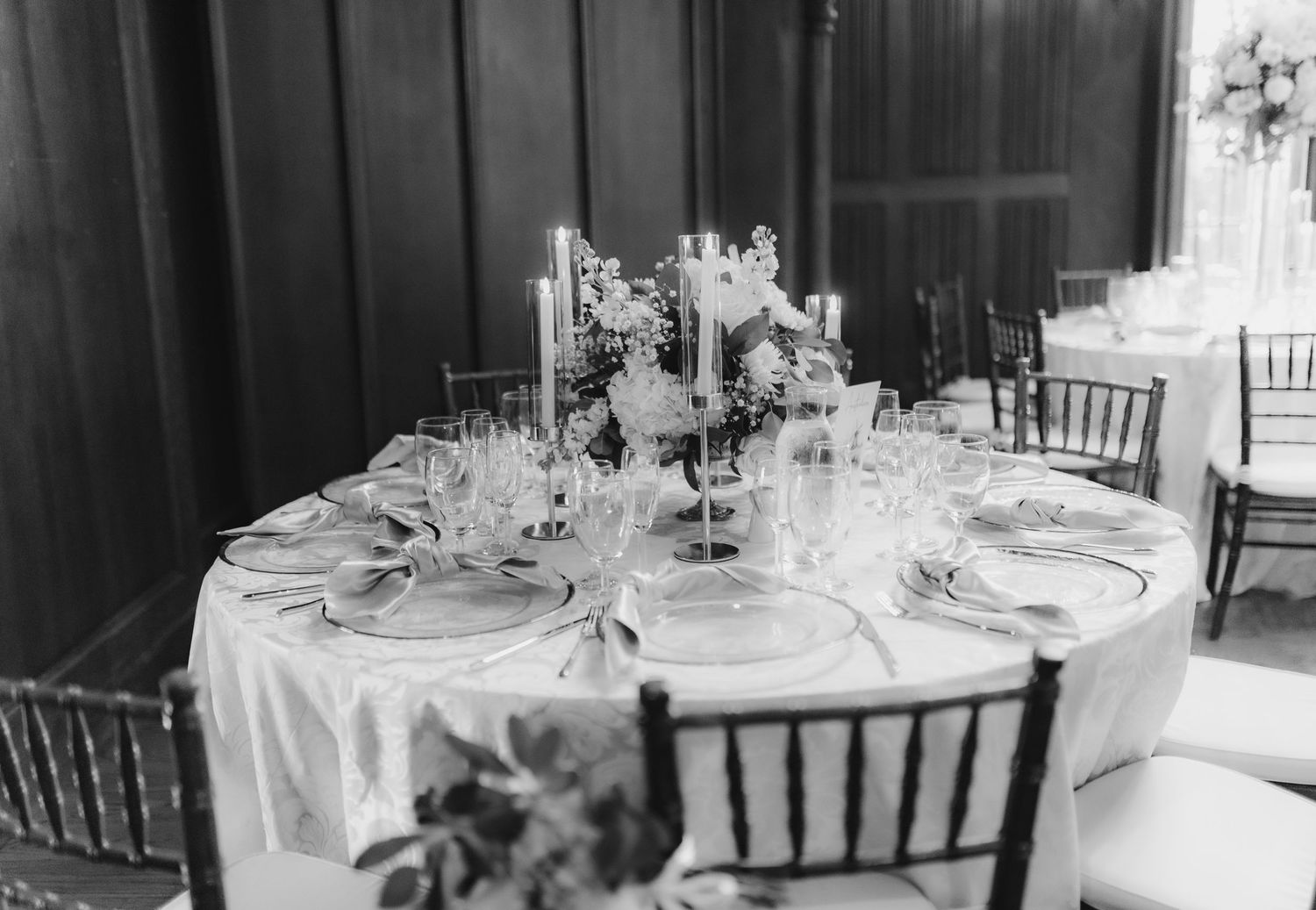 Black and white photo of elegantly set round reception table with candelabras and floral centerpiece.