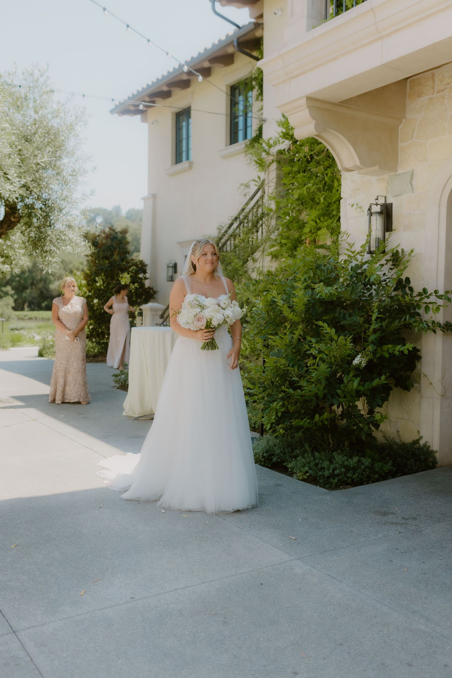 Bride in flowing white tulle gown holds white bouquet outside elegant stone wedding venue.