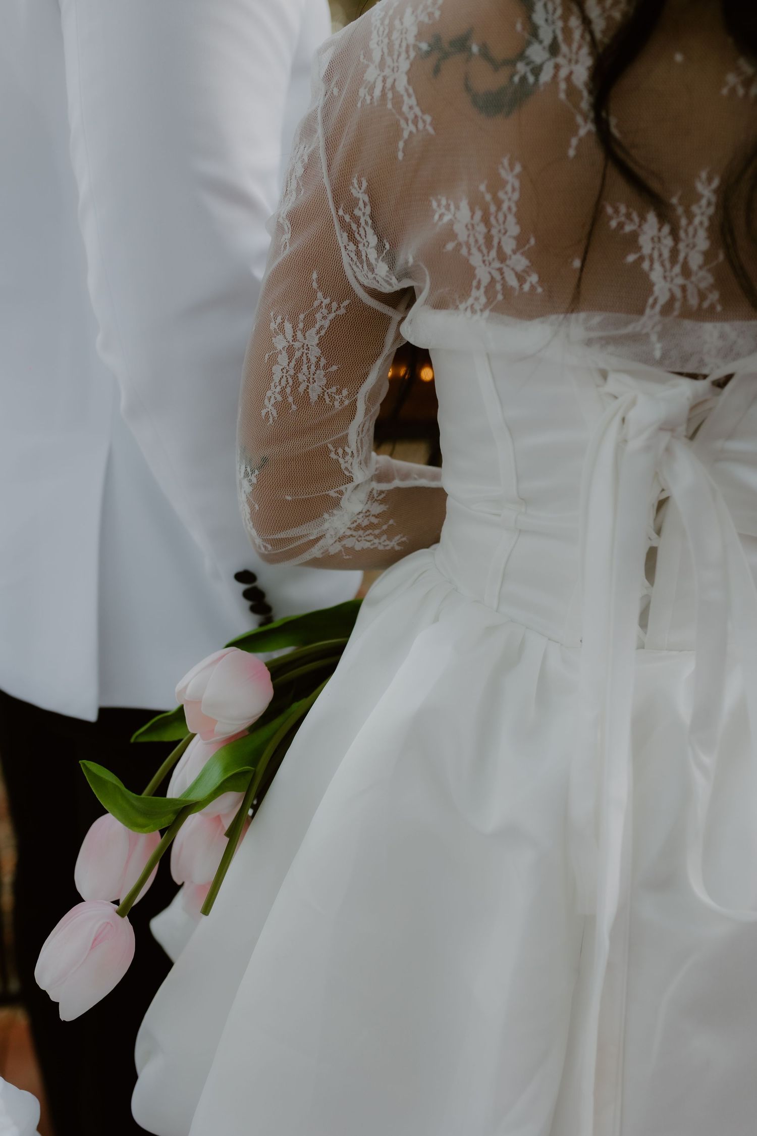 Close-up of bride in lace corset wedding gown holding pink tulips beside groom in white jacket.