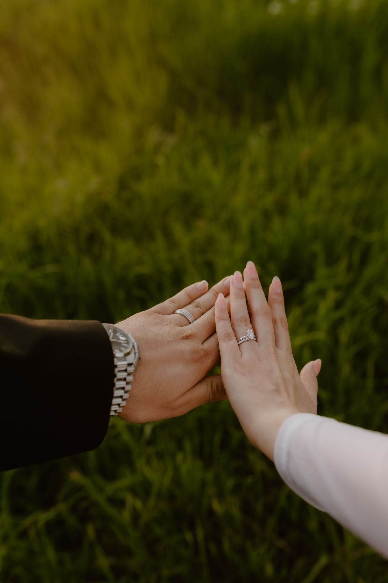 Close-up of a couple touching hands with wedding rings visible against a green grass background.