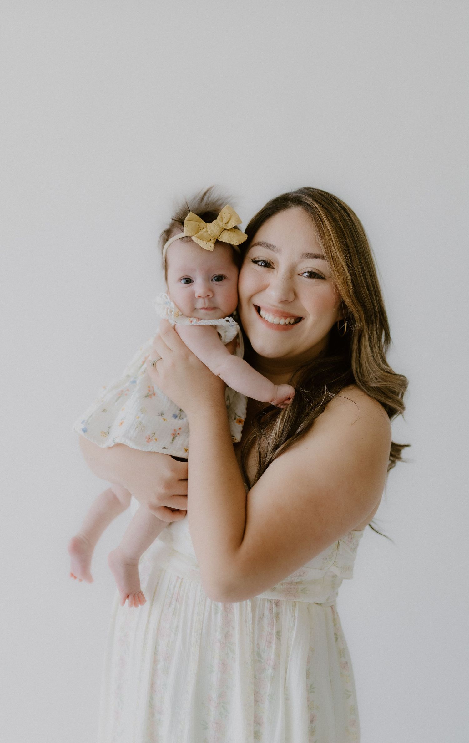 Smiling mother in white dress holding her baby girl in a floral outfit with a gold bow.