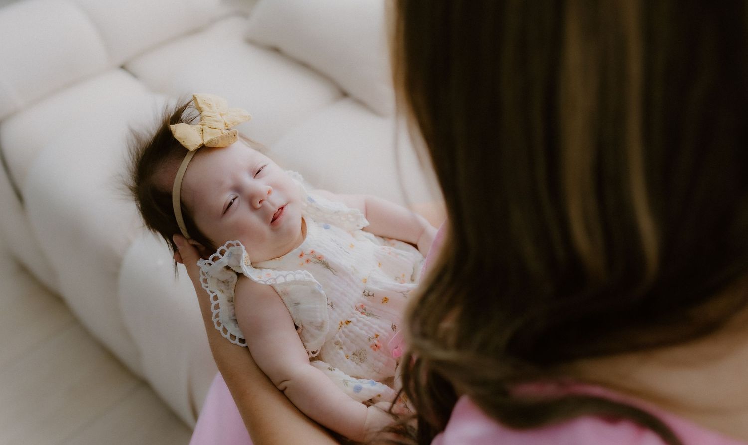 Mother in pink dress cradling her laughing baby girl wearing a yellow bow headband on a cream sofa.