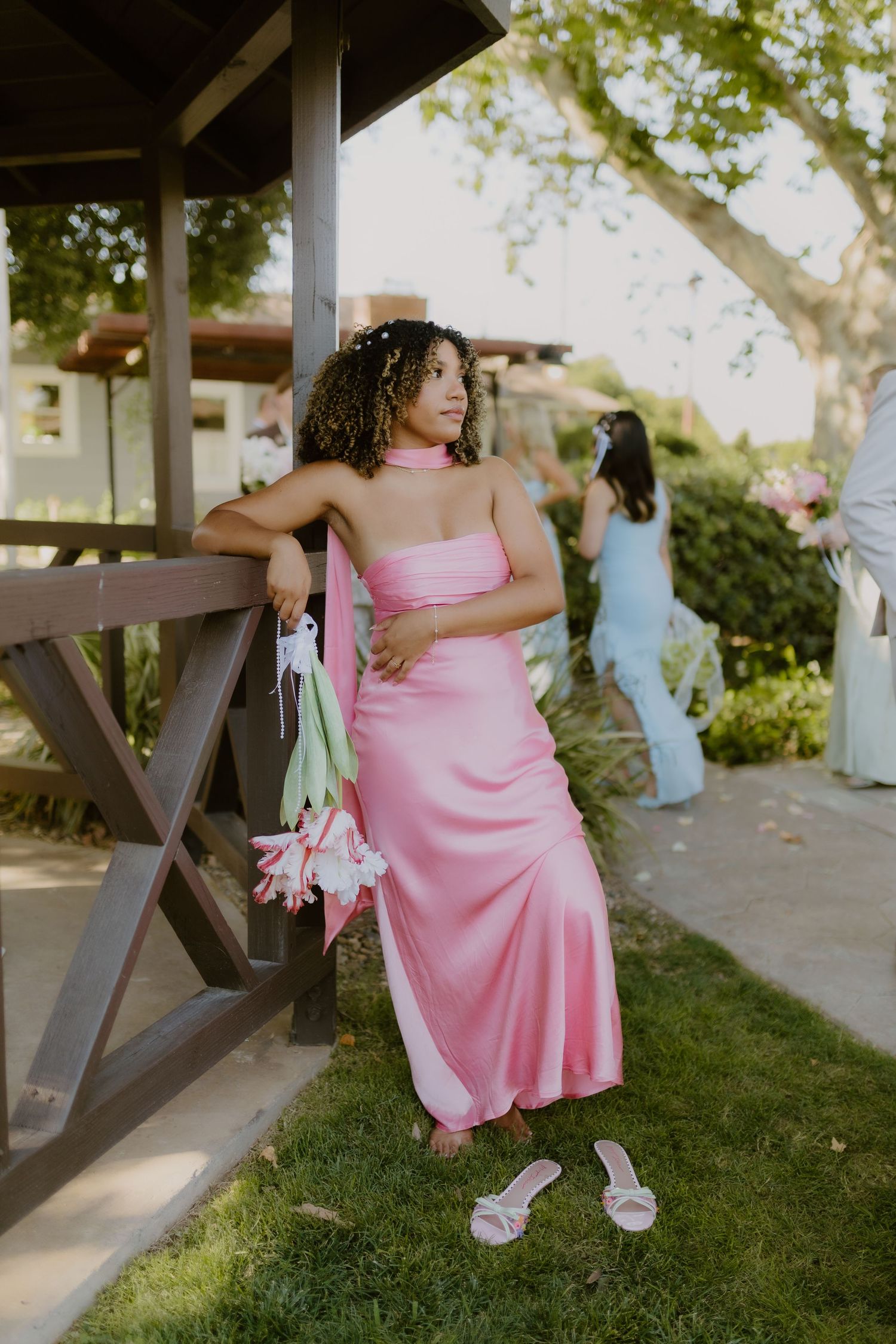 Bridesmaid in pink satin strapless gown holding tulips leaning against wooden gazebo post.