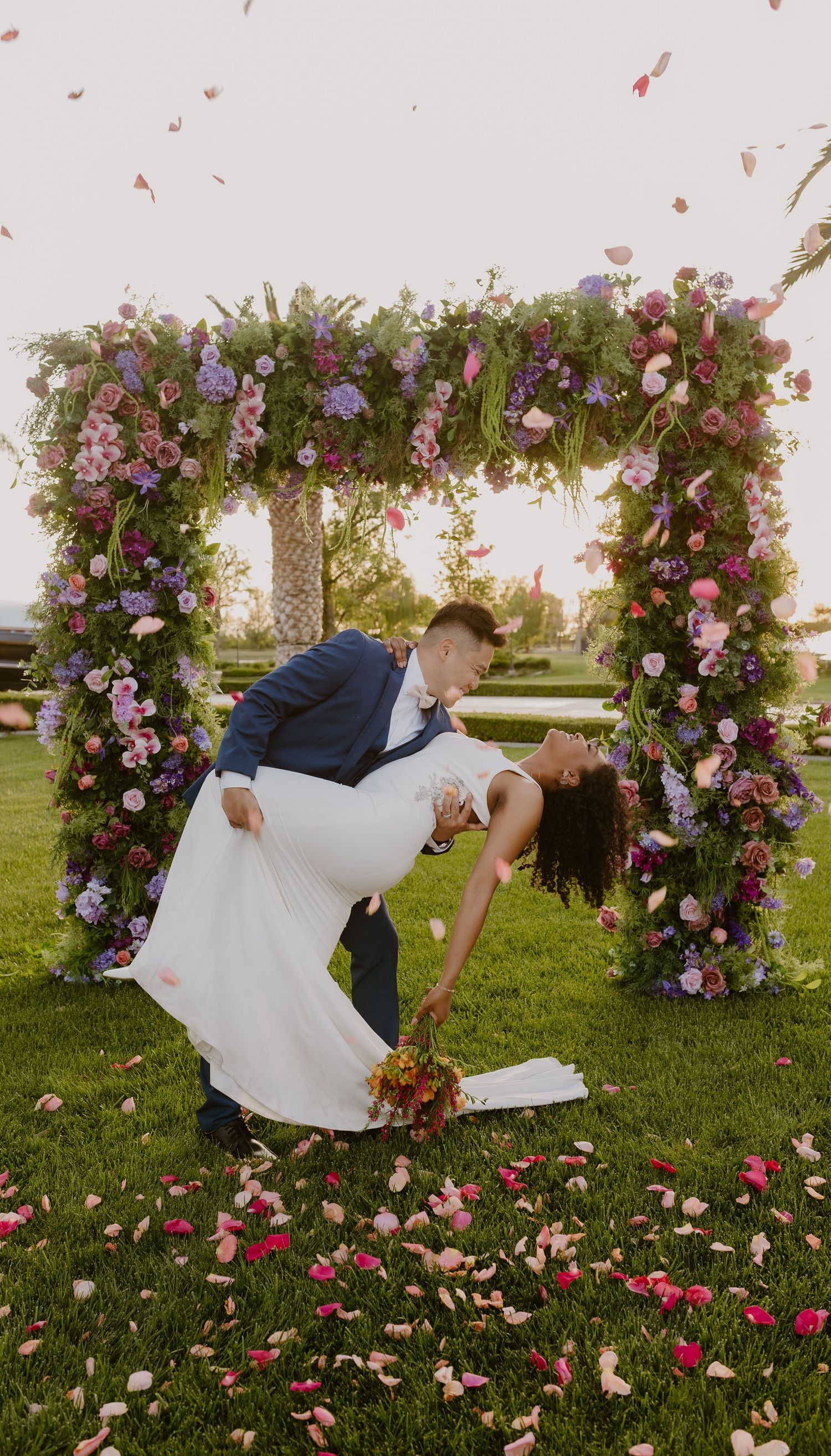 Groom dips bride under a stunning floral arch with pink and purple blooms as rose petals fall around them.