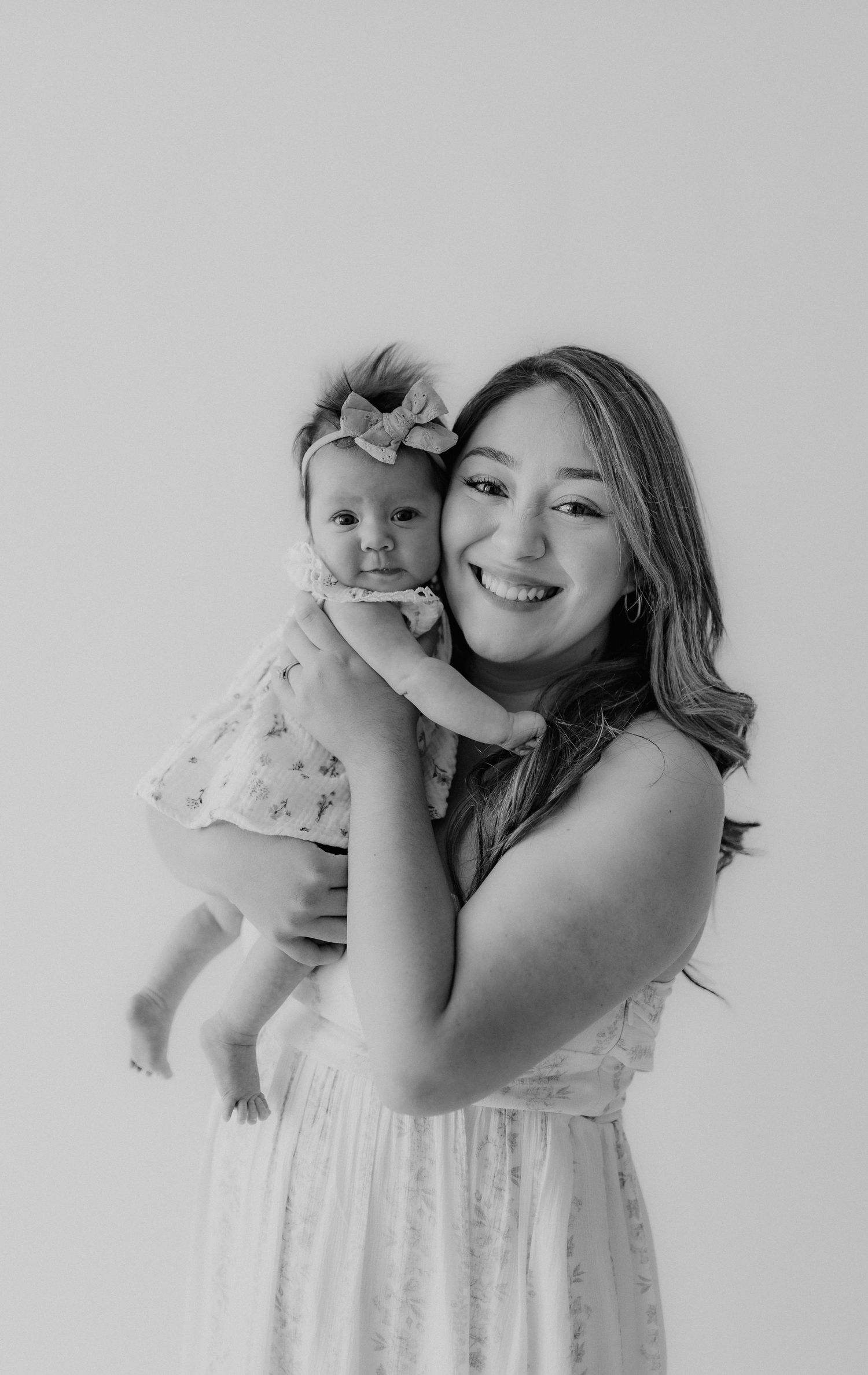 Black and white portrait of a smiling mother holding her baby girl with a bow headband.