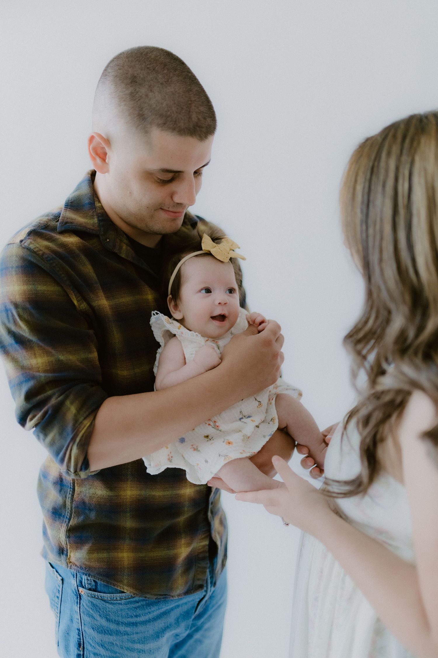 A father in a plaid shirt holds his infant daughter with a yellow bow as mother reaches out lovingly.
