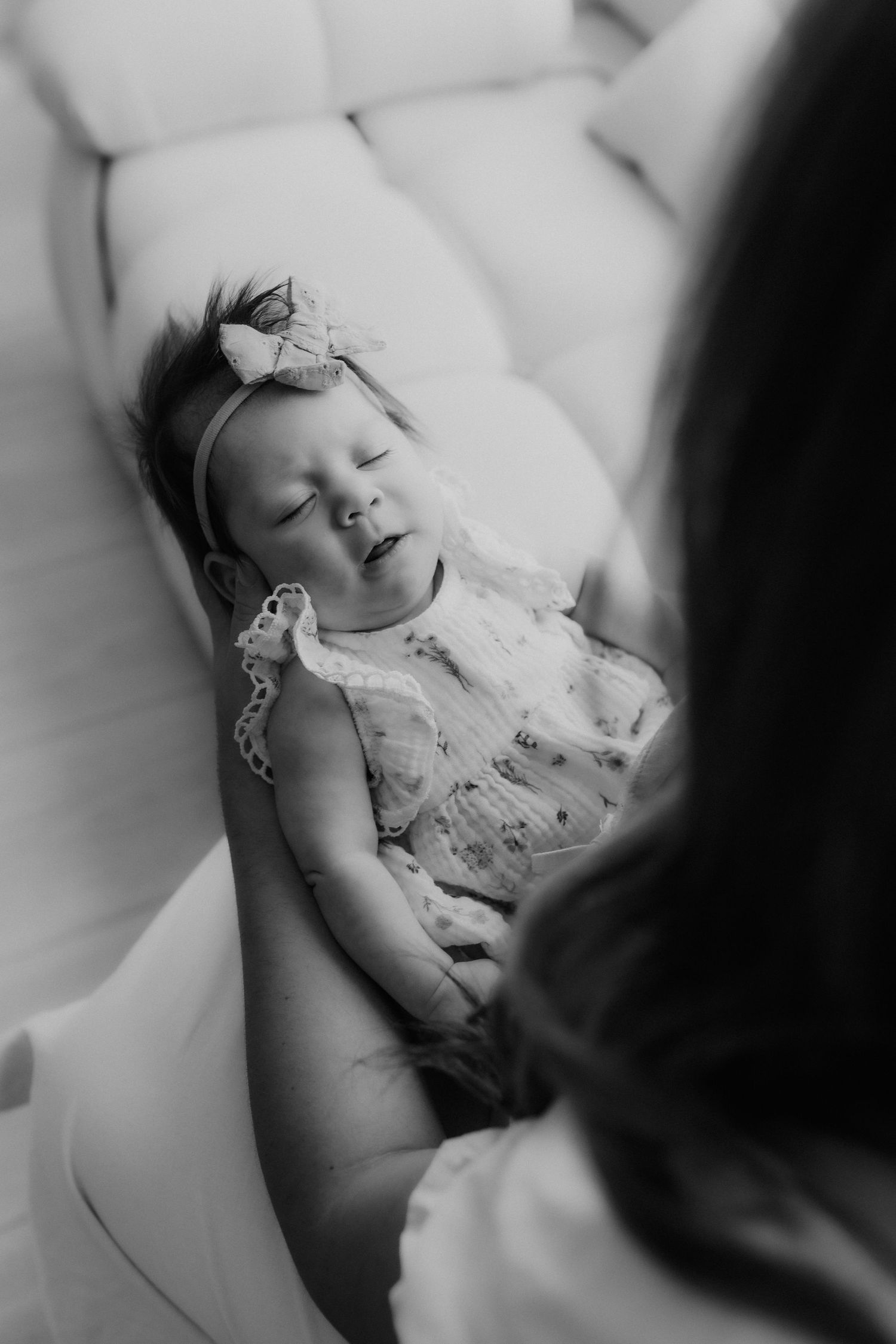 Black and white photo of a smiling baby girl in a bow headband resting in her mothers arms on a sofa.