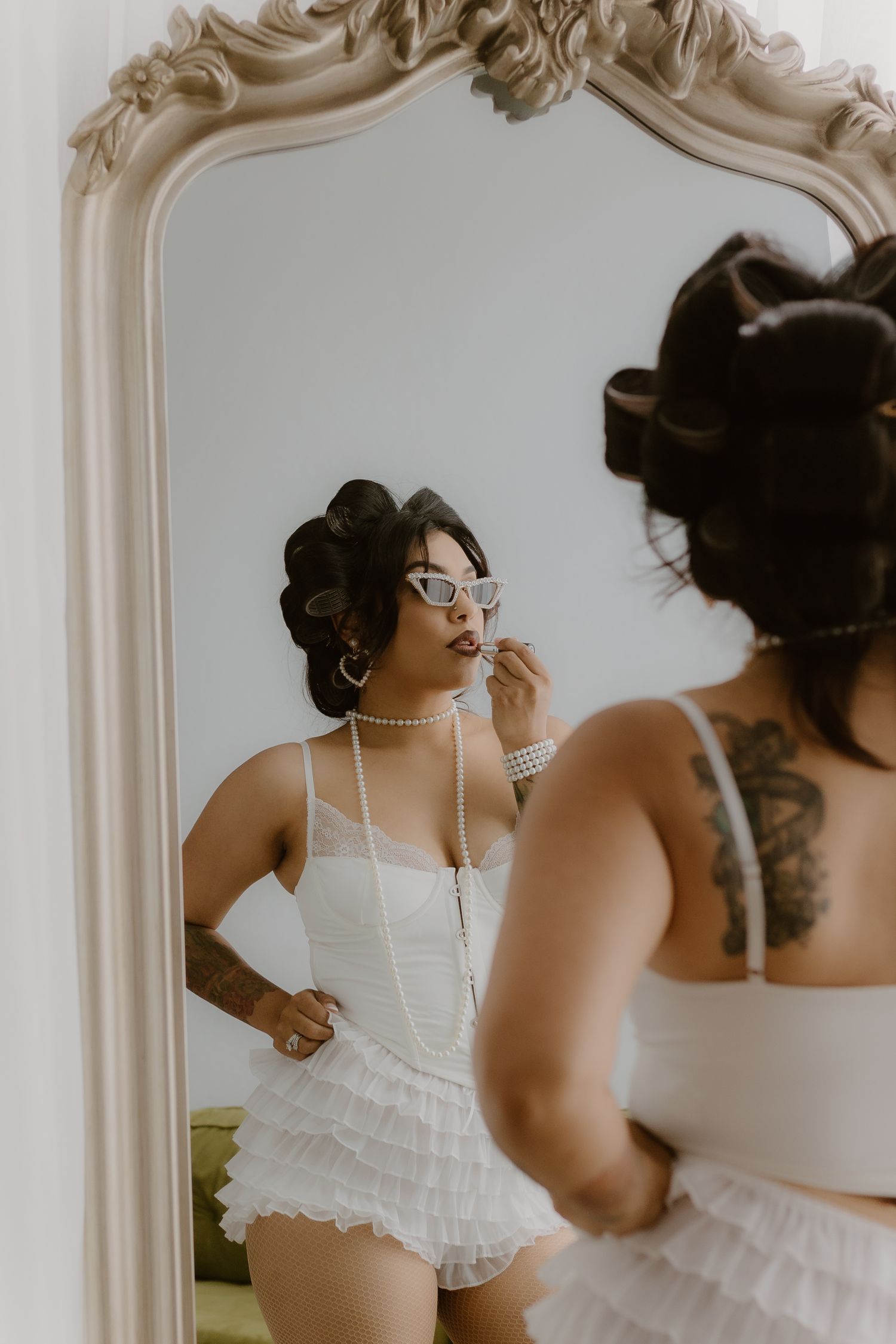 Woman with hair rollers in white ruffled corset set applying lipstick while looking in ornate mirror. Tulare, California