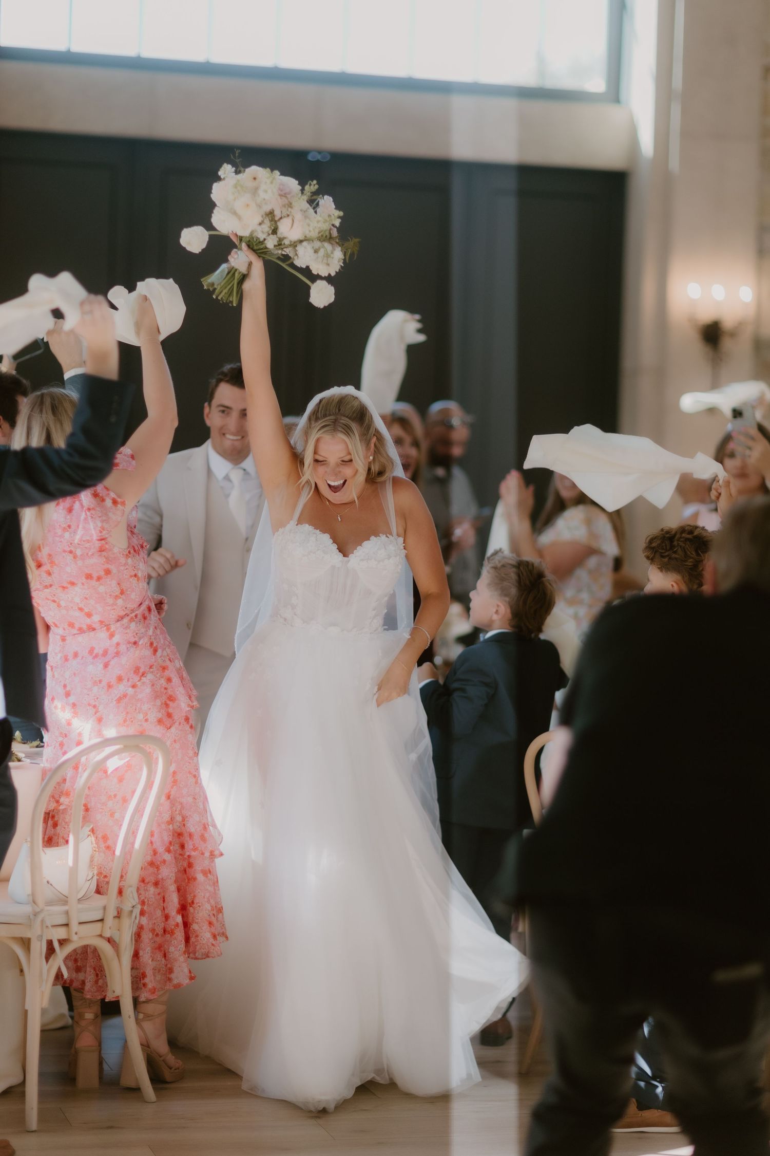 Excited bride raises bouquet triumphantly while entering wedding reception as guests wave and celebrate.