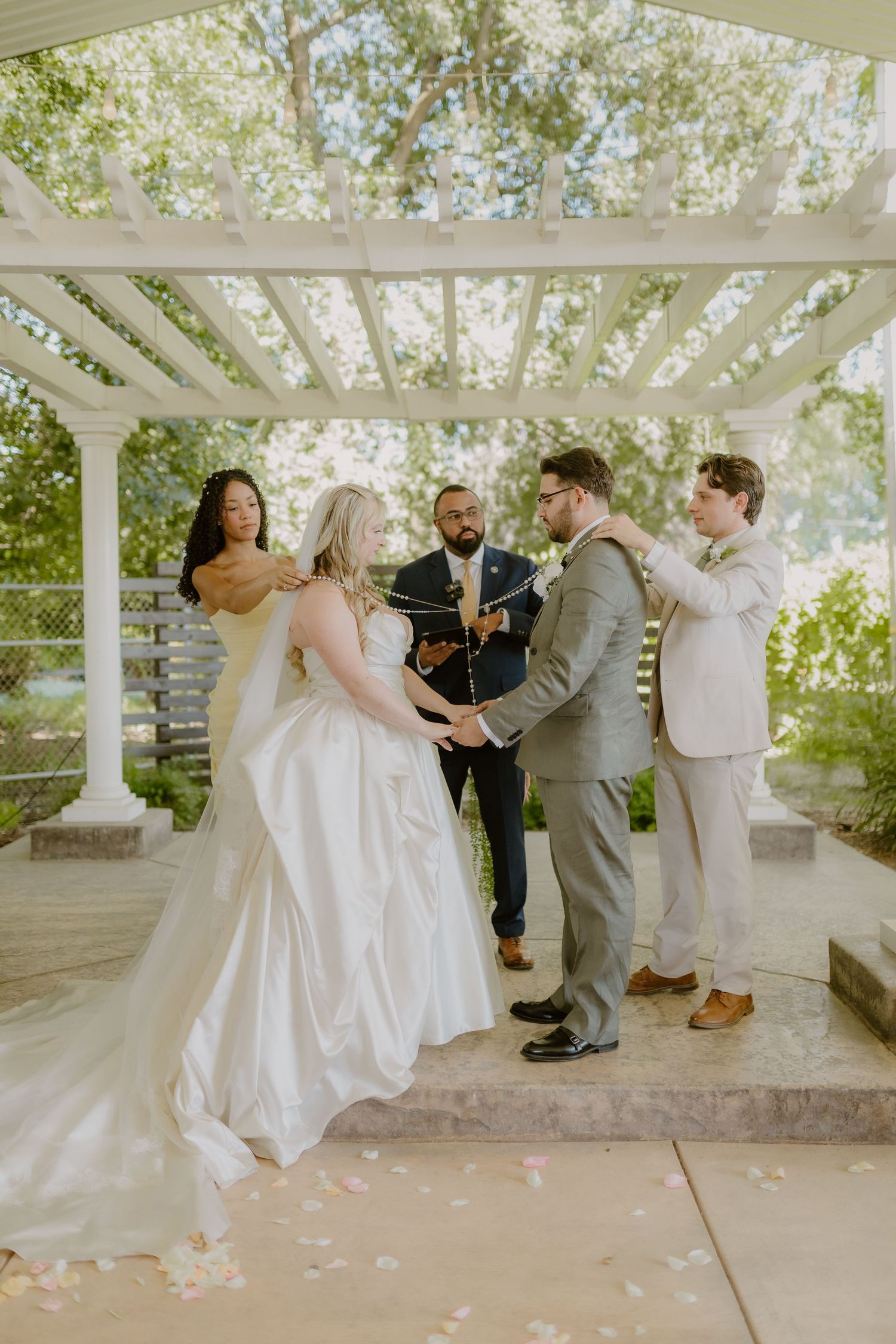 Wedding party performing hand-fasting ritual under white pergola with officiant and best man.