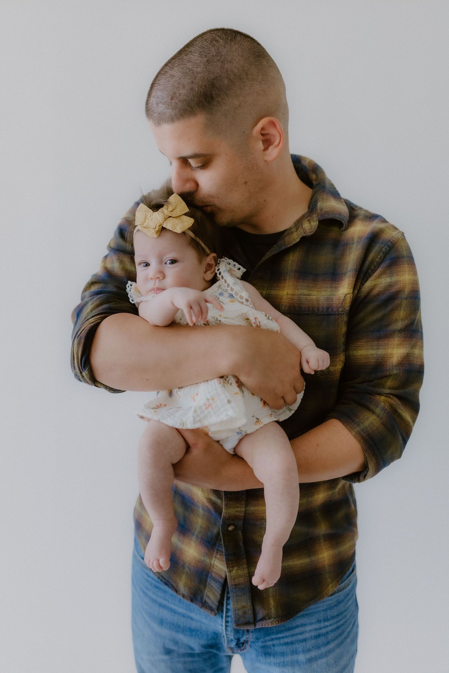 Father in plaid shirt tenderly kissing his baby girl in a lace outfit with a yellow bow headband.
