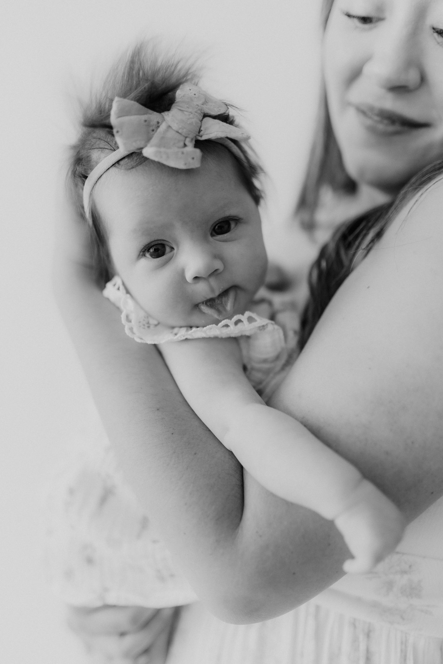 Black and white close-up of a newborn baby girl with a bow headband sticking out her tongue in mothers arms.