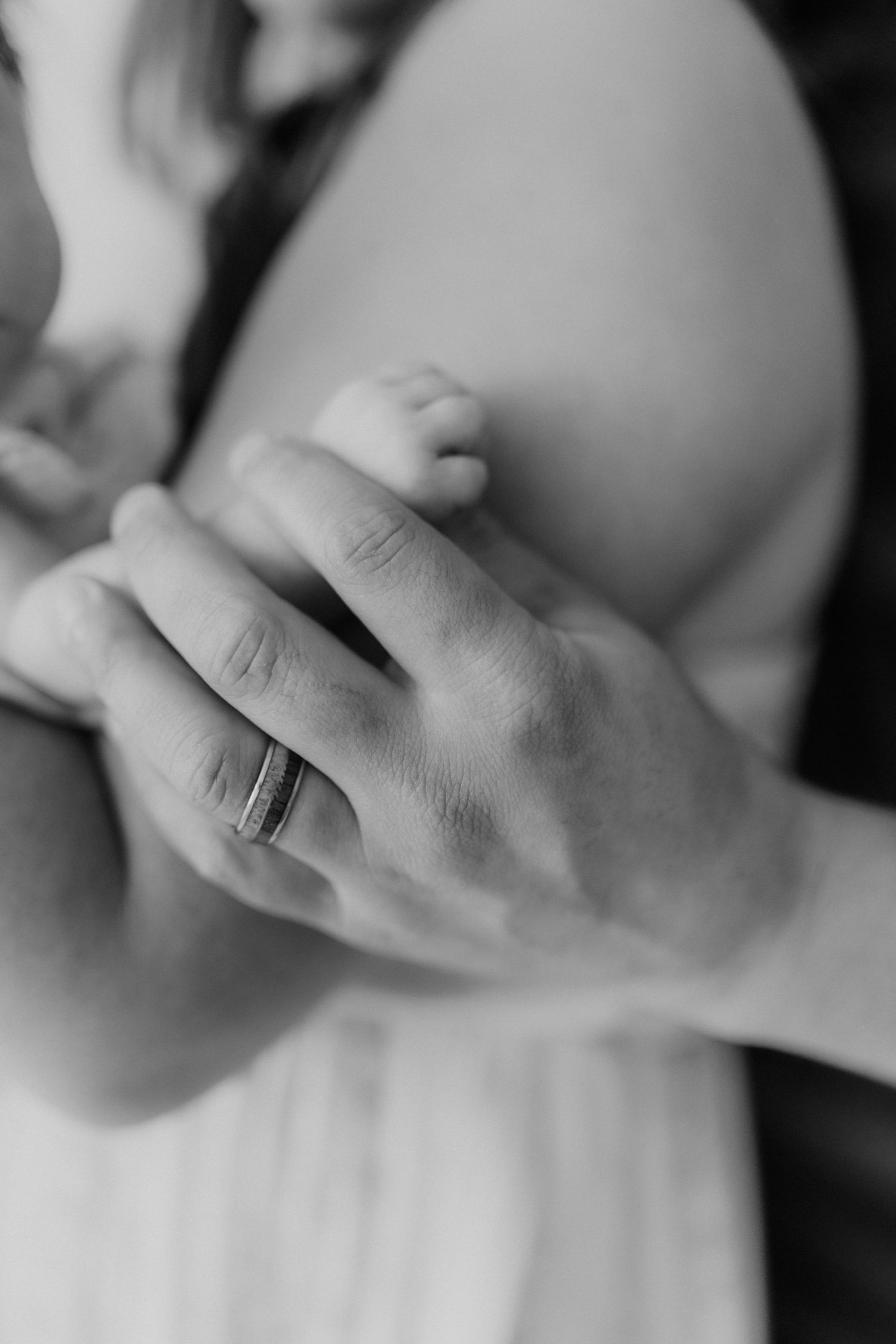 Black and white close-up of a fathers hand with wedding band gently touching his newborns tiny foot.