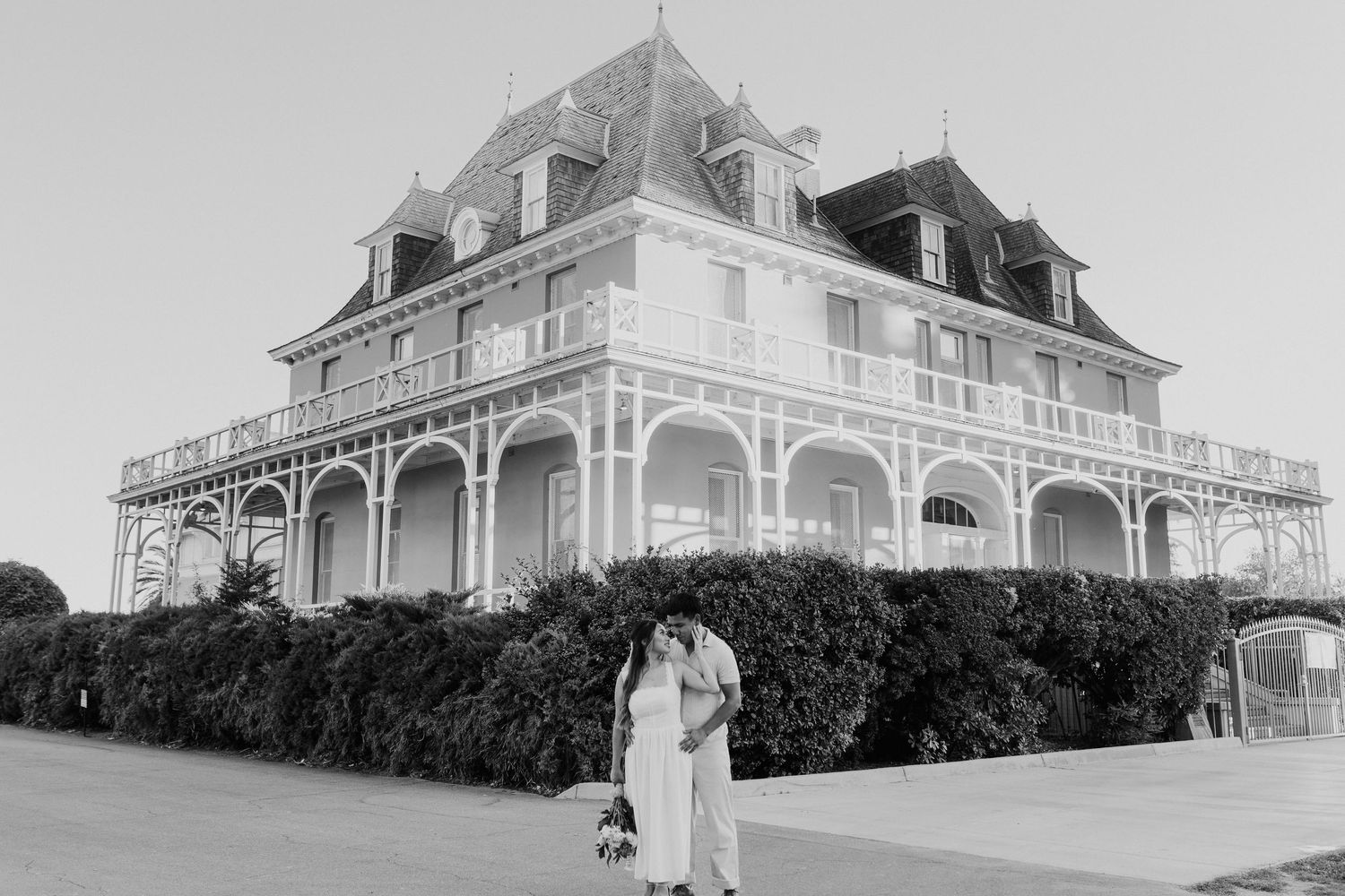 Black and white engagement photo of couple embracing before a large ornate Victorian-style building.