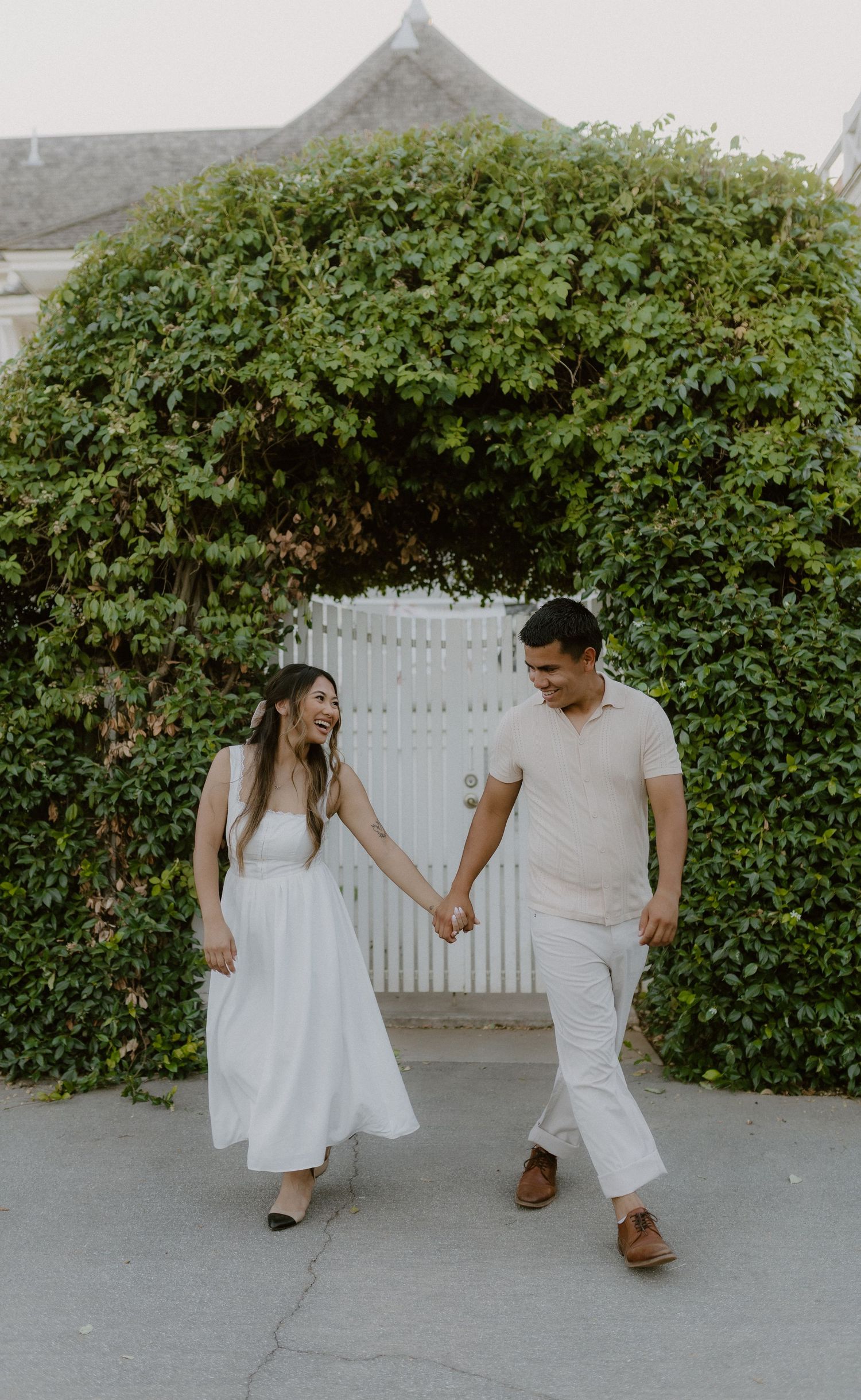 Smiling couple in white outfits holding hands walking near ivy-covered white gate.